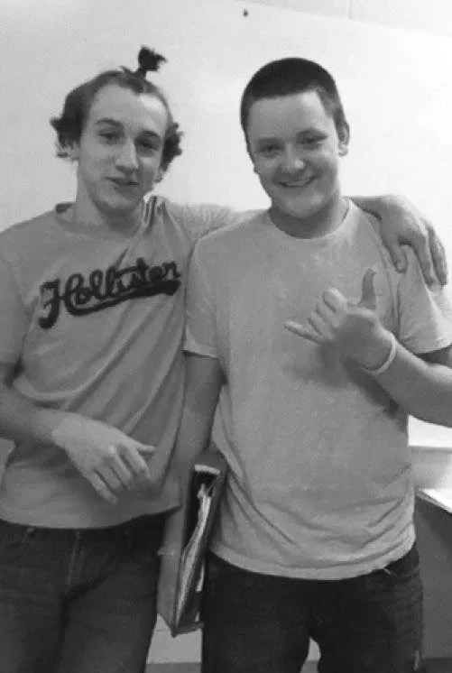 Two young men standing together, smiling indoors in front of a plain wall.