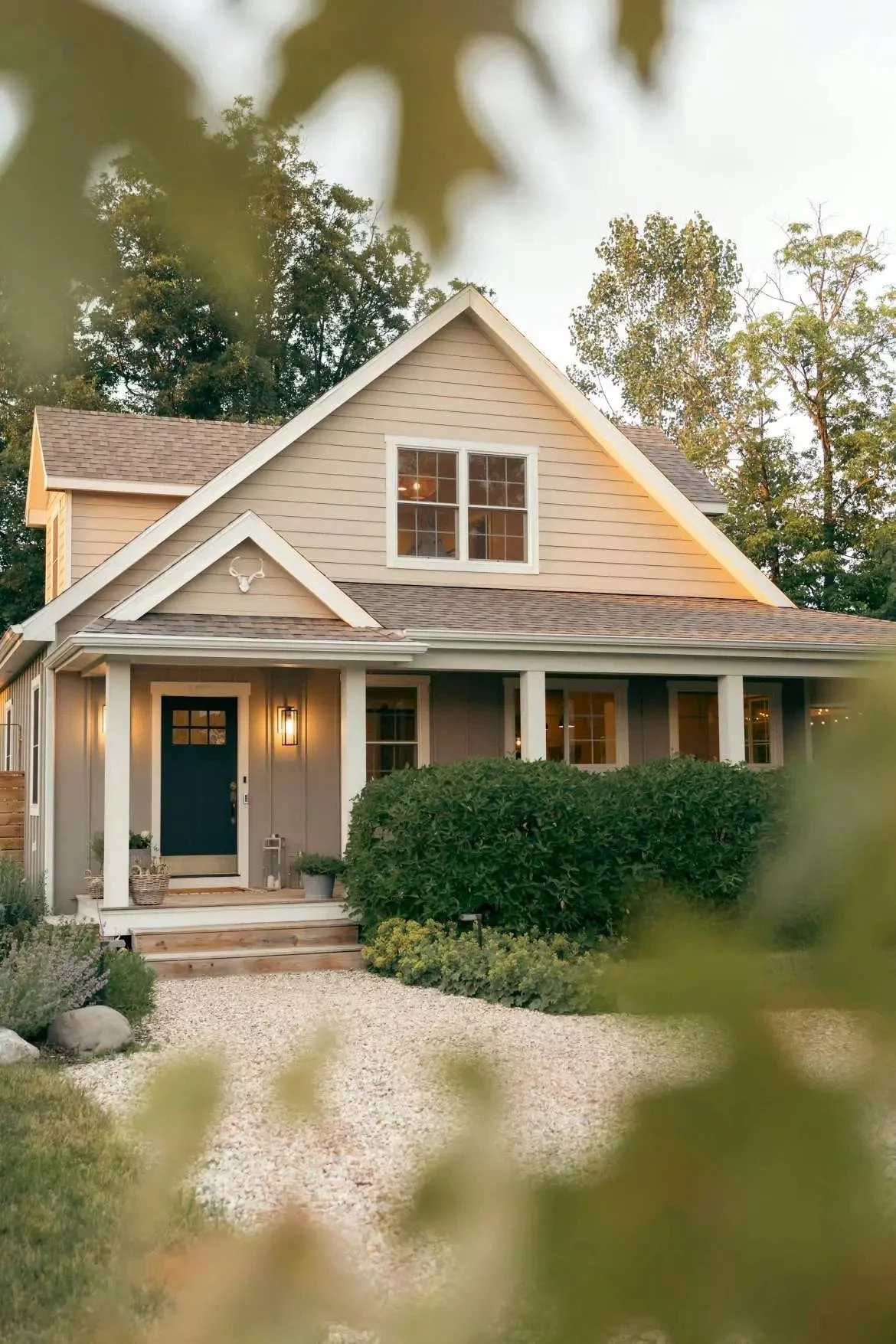 A two-story house with beige siding, white trim, and a dark blue front door, surrounded by greenery and trees, with a gravel pathway leading to the front steps.