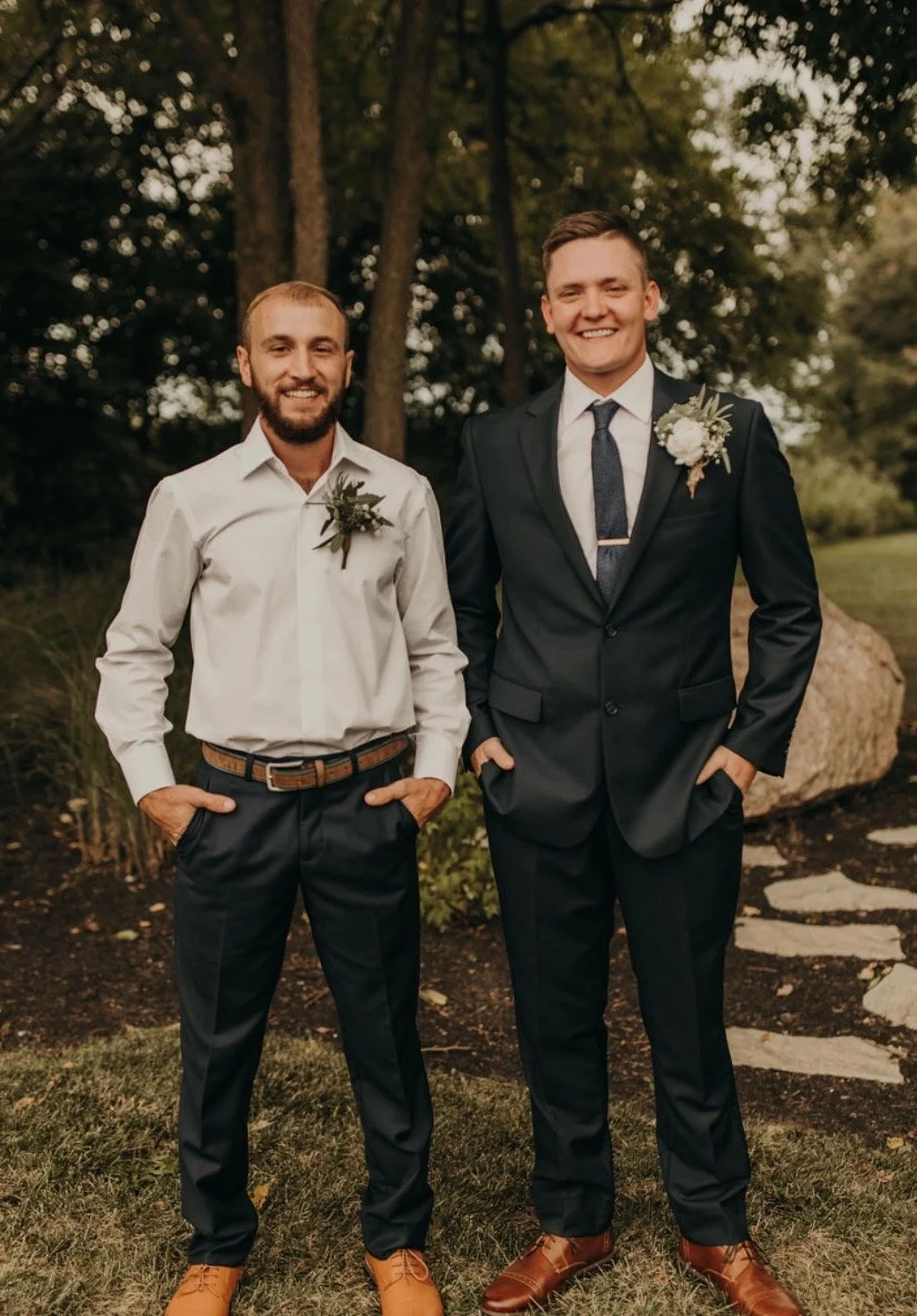 Two men dressed in formal attire standing outdoors with trees in the background. One is wearing a white shirt and the other a navy suit.