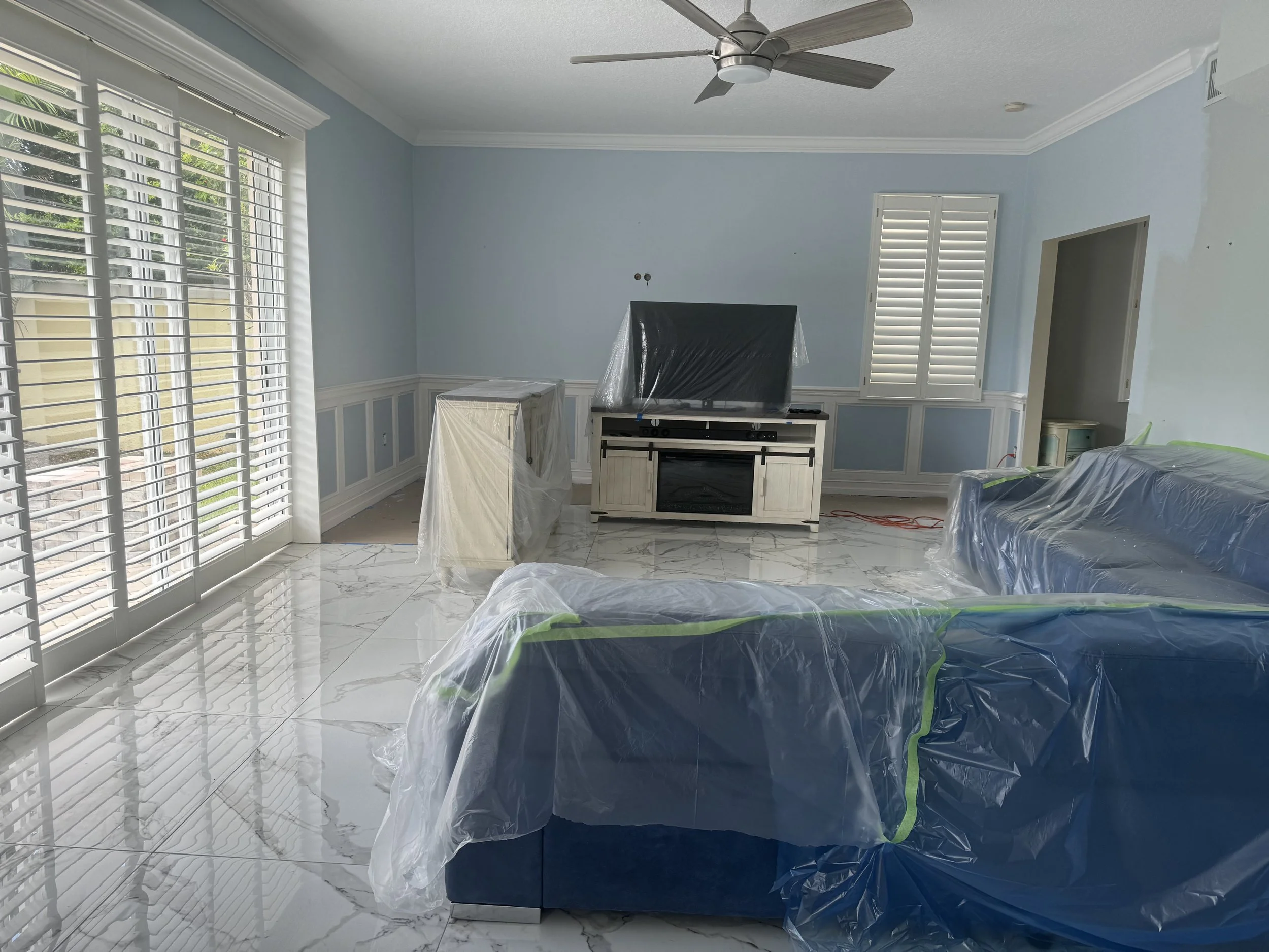 Living room with large window shutters, a ceiling fan, and furniture covered in plastic, including a sofa and a TV stand.