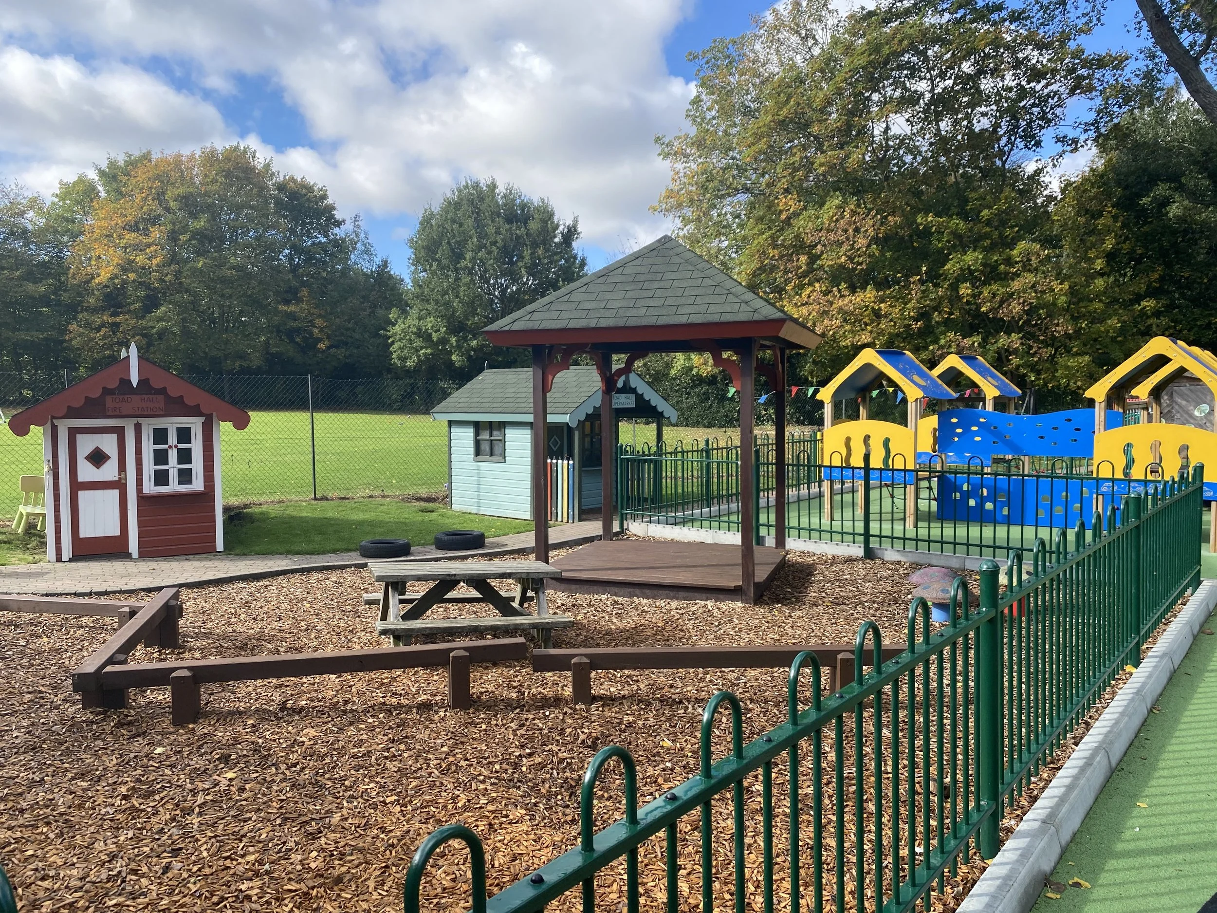 Kids' playground with small playhouse, picnic table, and colorful climbing structures, surrounded by a green fence and trees in the background under partly cloudy sky.