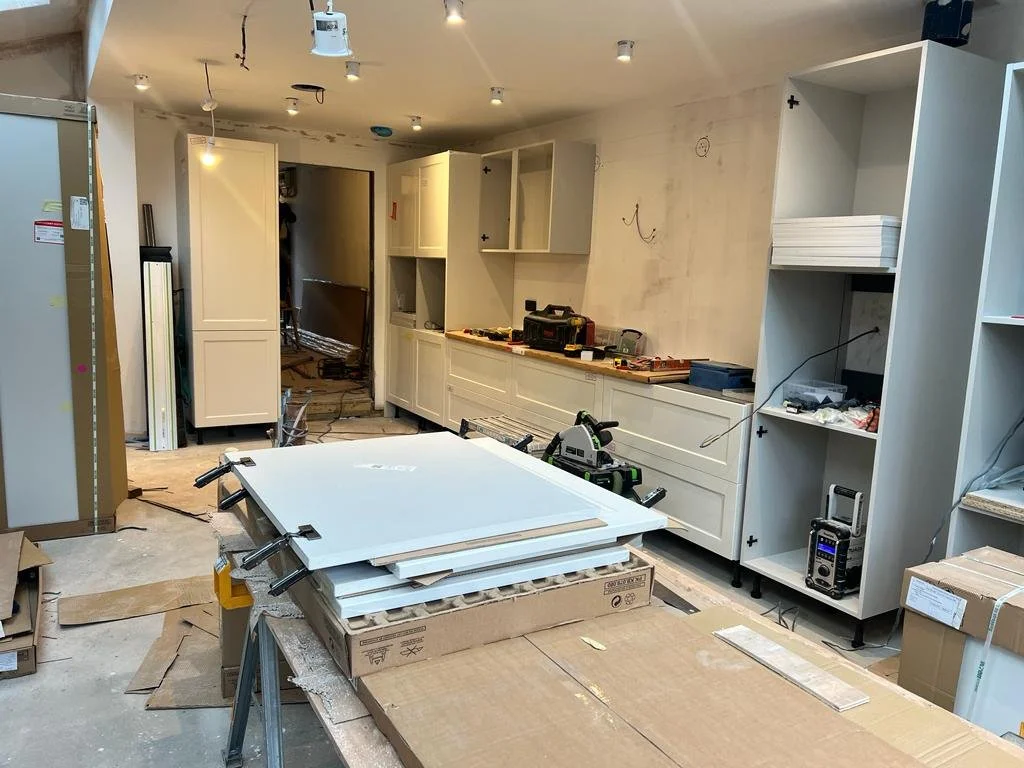 A room under construction with partially installed white shelves, a work table with tools, and cardboard boxes, indicating ongoing furniture or cabinetry installation.
