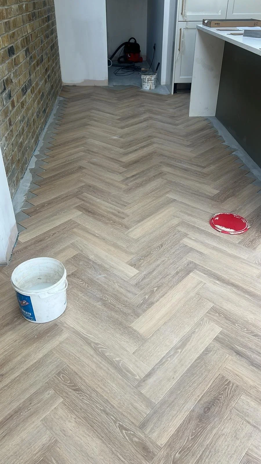 A room with a herringbone wood floor installation in progress, with a white bucket and a container of red paint on the floor, and construction tools and supplies visible in the background.