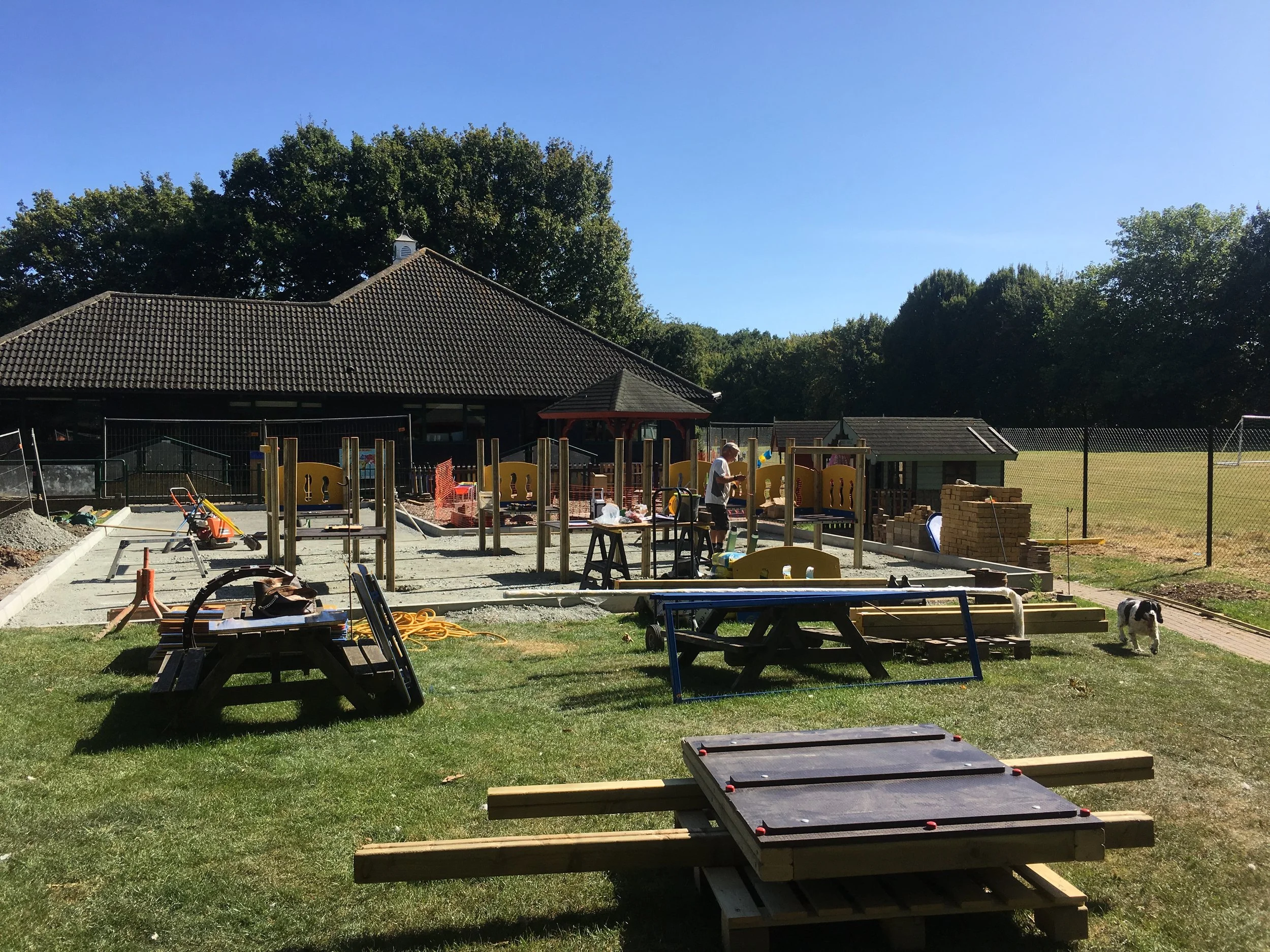 Construction site of a playground with workers, scattered tools, and building materials, with a dog on the grass and trees in the background.