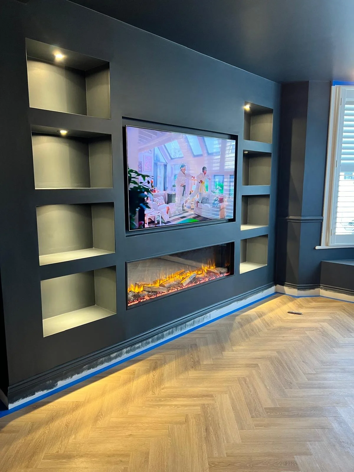 Modern living room featuring a dark wall with built-in display shelves, a flat-screen TV, and a linear electric fireplace beneath it. The room has wood flooring and a window with white blinds.