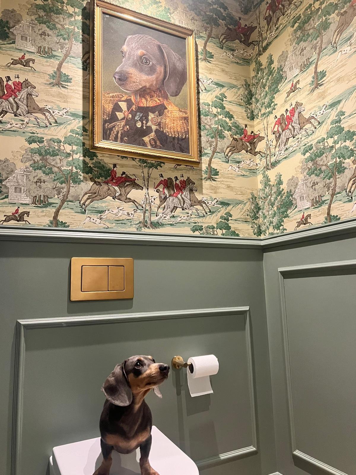 A small dog sitting on a white platform in a room with green walls and decorative molding. Behind the dog, there is a toilet paper holder with a roll of toilet paper and a gold-colored flush panel on the wall. The upper section of the wall features a large framed portrait of a dog in a military uniform, hanging above a mural of people riding horses and dogs running through a landscape.