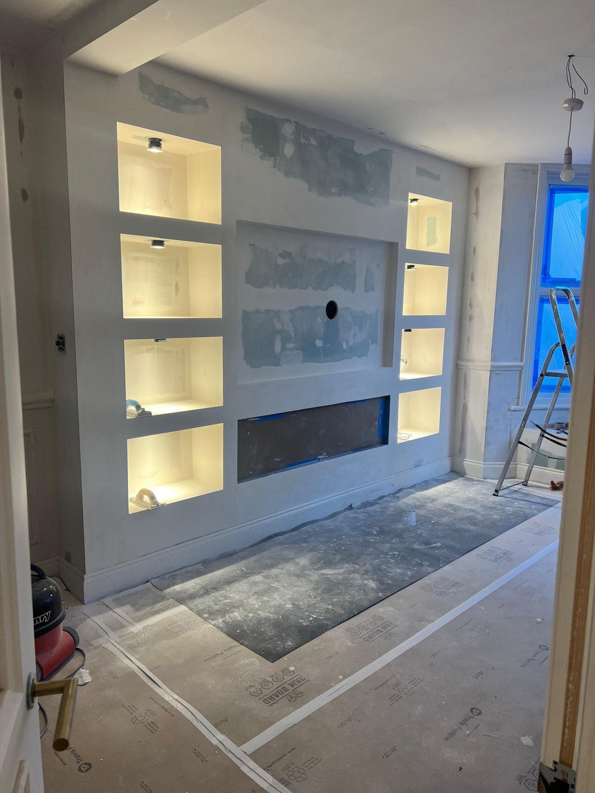 A room under renovation with a built-in entertainment wall with recessed shelves, some lights installed, and a hole for a TV. The floor is covered with protective paper and an area rug, and a ladder is present near a window with blue protective film. Tools and equipment are scattered around.