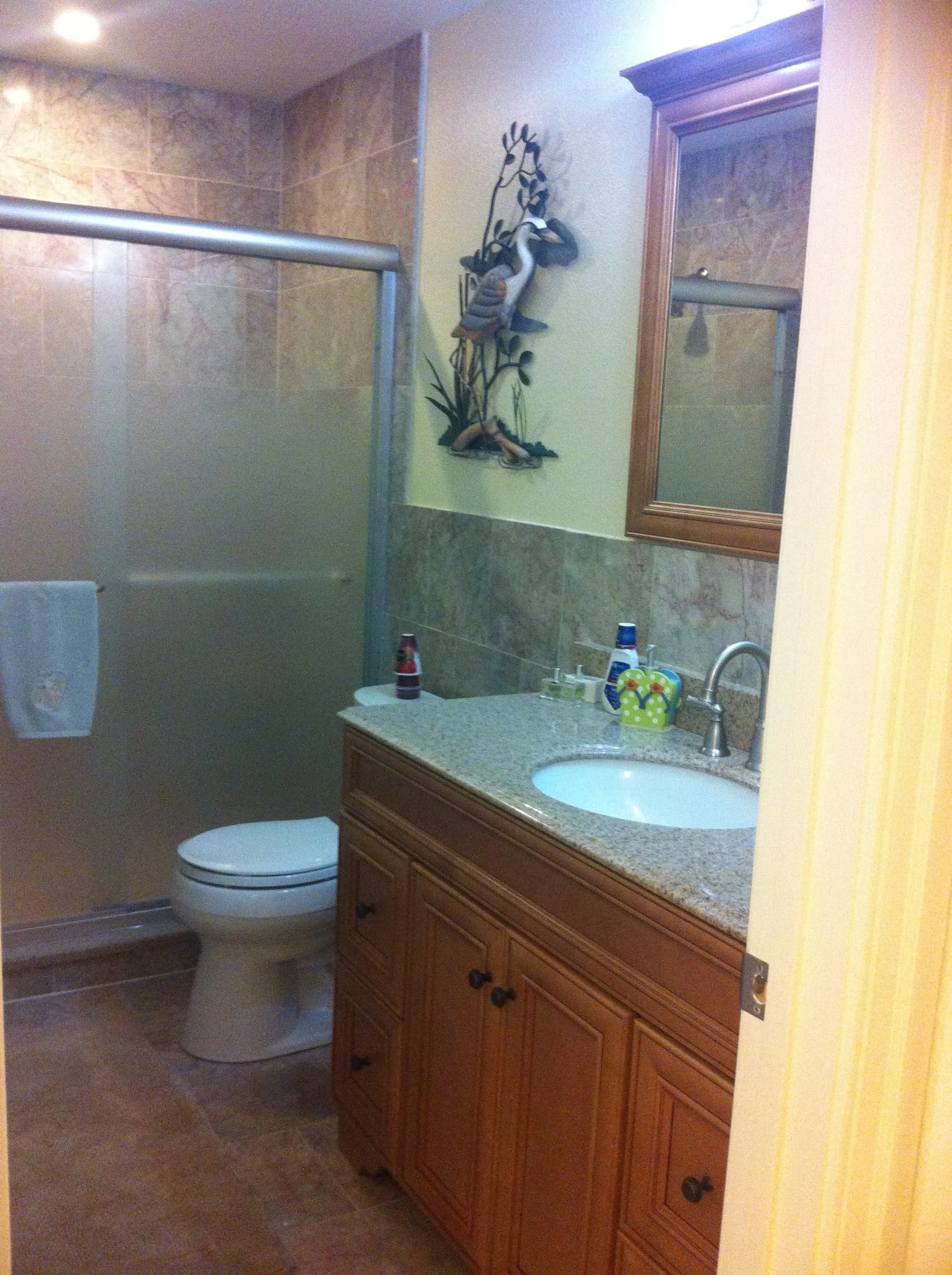 A bathroom with wooden vanity, granite countertop, mirror, shower with frosted glass door, and fish wall decoration.