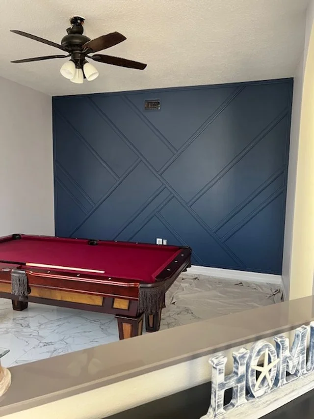 A room with a blue accent wall featuring geometric patterns, a red pool table with tassels on the corners, a white marble floor, a ceiling fan with lights, and a decorative sign that says "HOME" in the foreground.