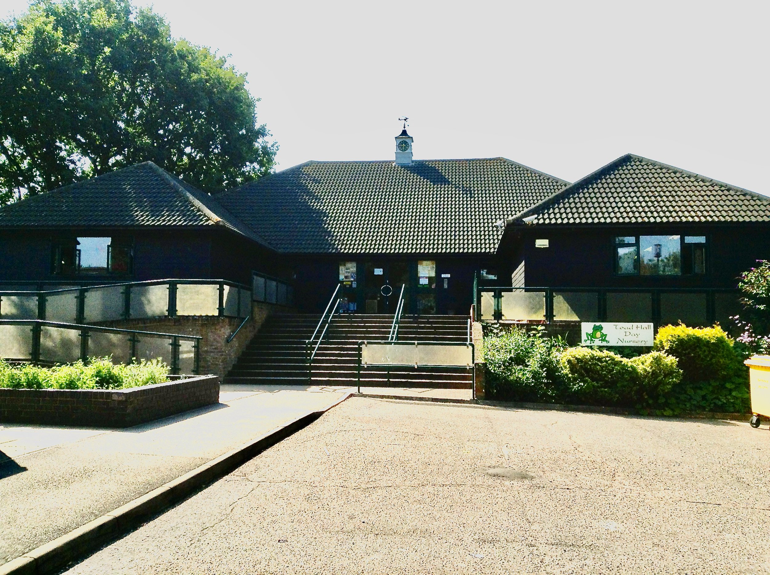 Front view of Toad Hall Day Nursery with stairs leading up to the entrance, surrounded by greenery and bushes, under a clear sky.