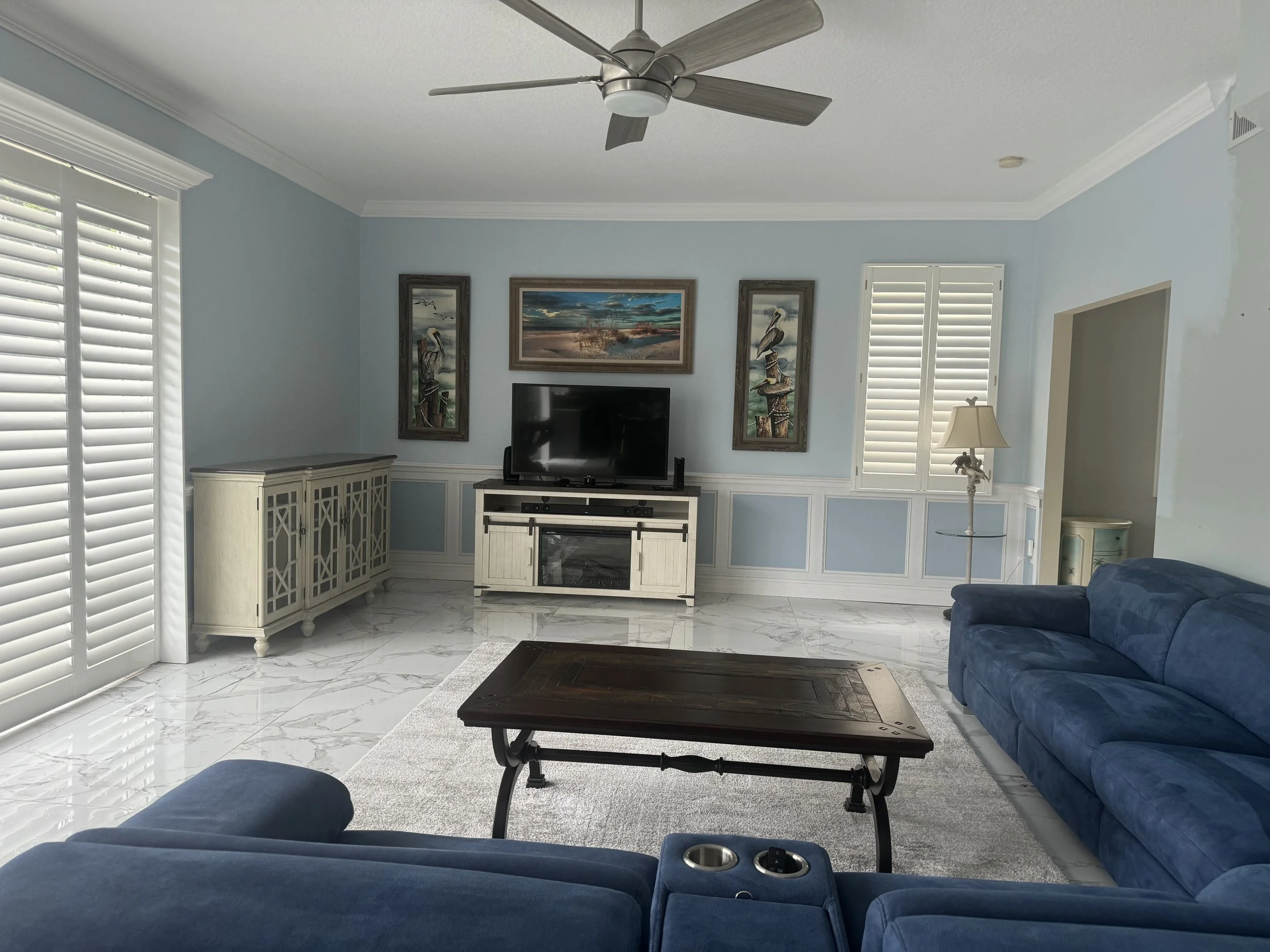 Living room with light blue walls, white trim, white marble floor, blue sofas, a wooden coffee table, a TV on a white entertainment stand, framed pictures of pelicans and a beach scene on the wall, a floor lamp near a window with white shutters, a white sideboard, a ceiling fan, and a light-colored area rug.