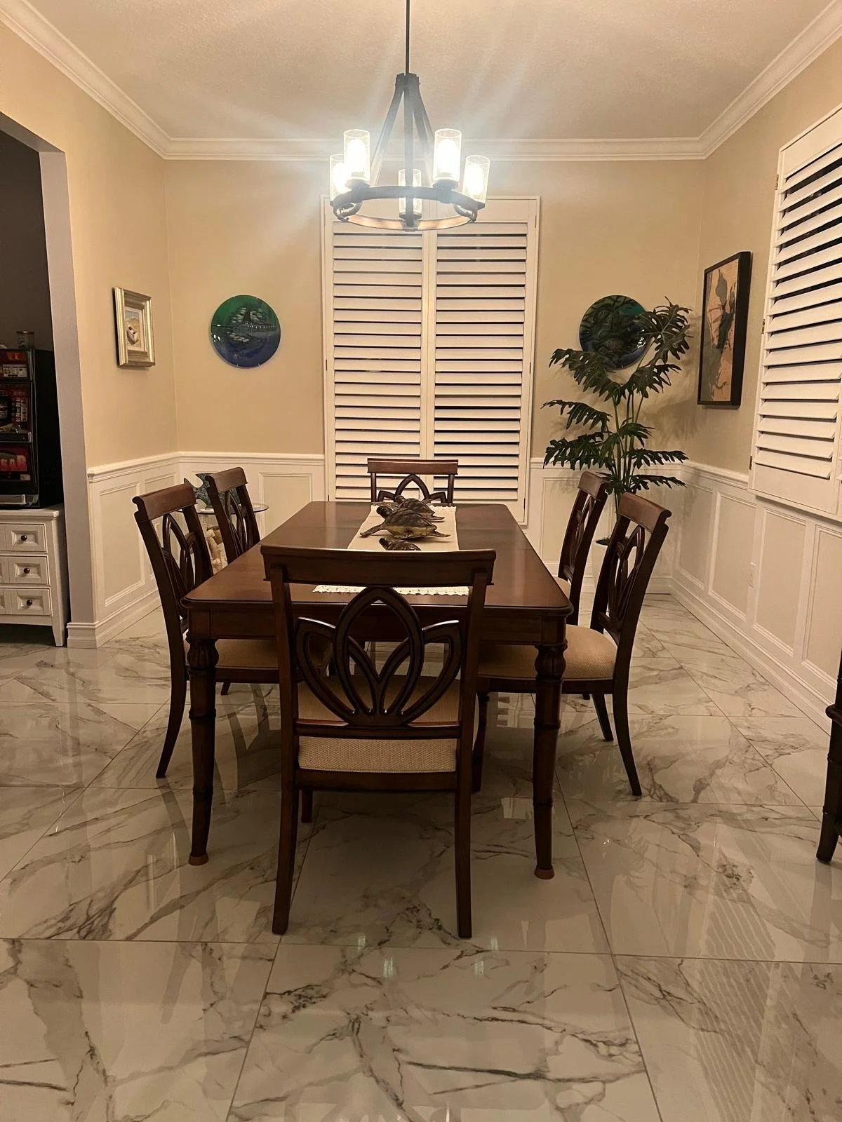 A dining room with a wooden table and six chairs, marble tile flooring, a chandelier, window shutters, a large plant, and artwork on the walls.