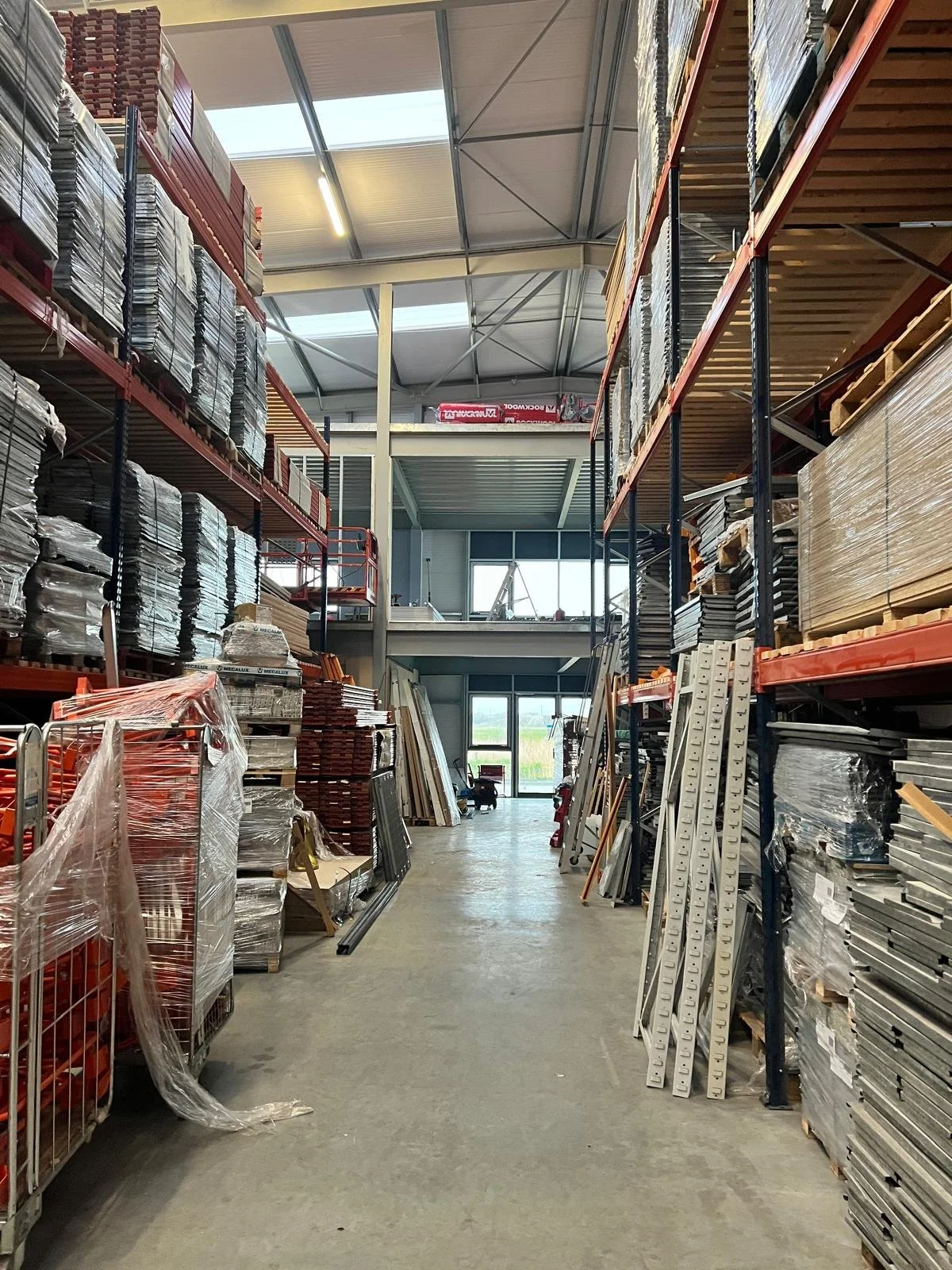 Interior of a warehouse displaying shelves stocked with building materials, including metal and wood, with a clear aisle leading to a glass door at the back.