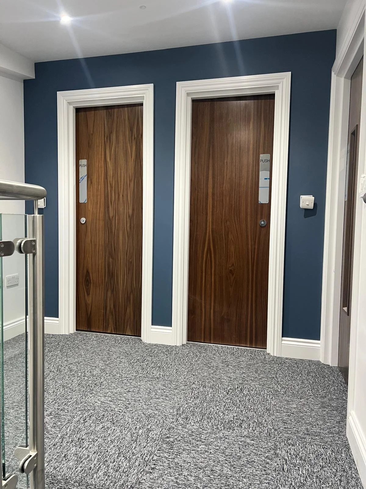 Two wooden elevator doors with push labels, framed by white decorative trim, against a dark blue wall and gray carpeted floor.