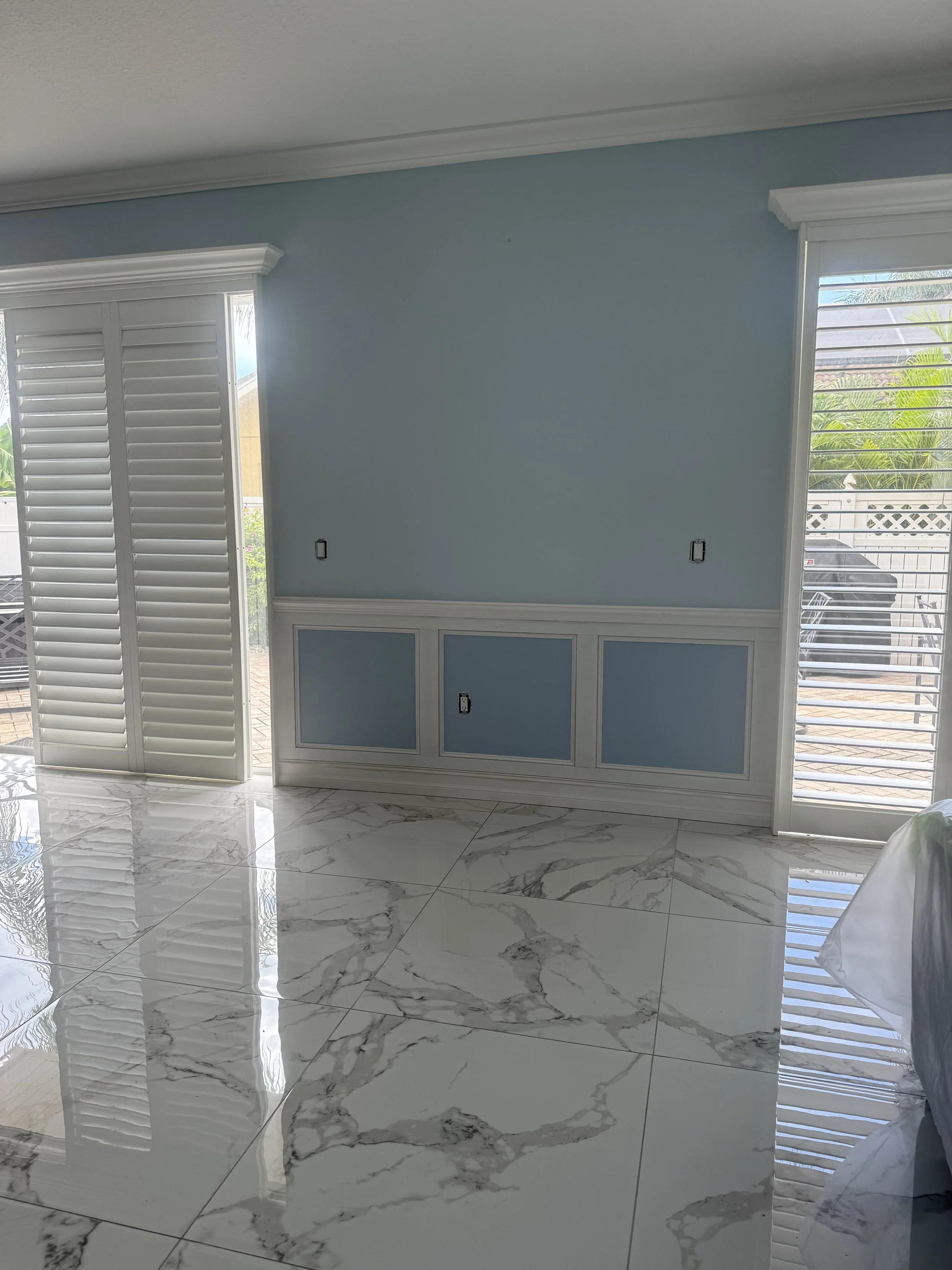 Empty room with light blue walls, white crown molding, and marble tile flooring with gray veining. Two large windows with white shutters letting in natural light.