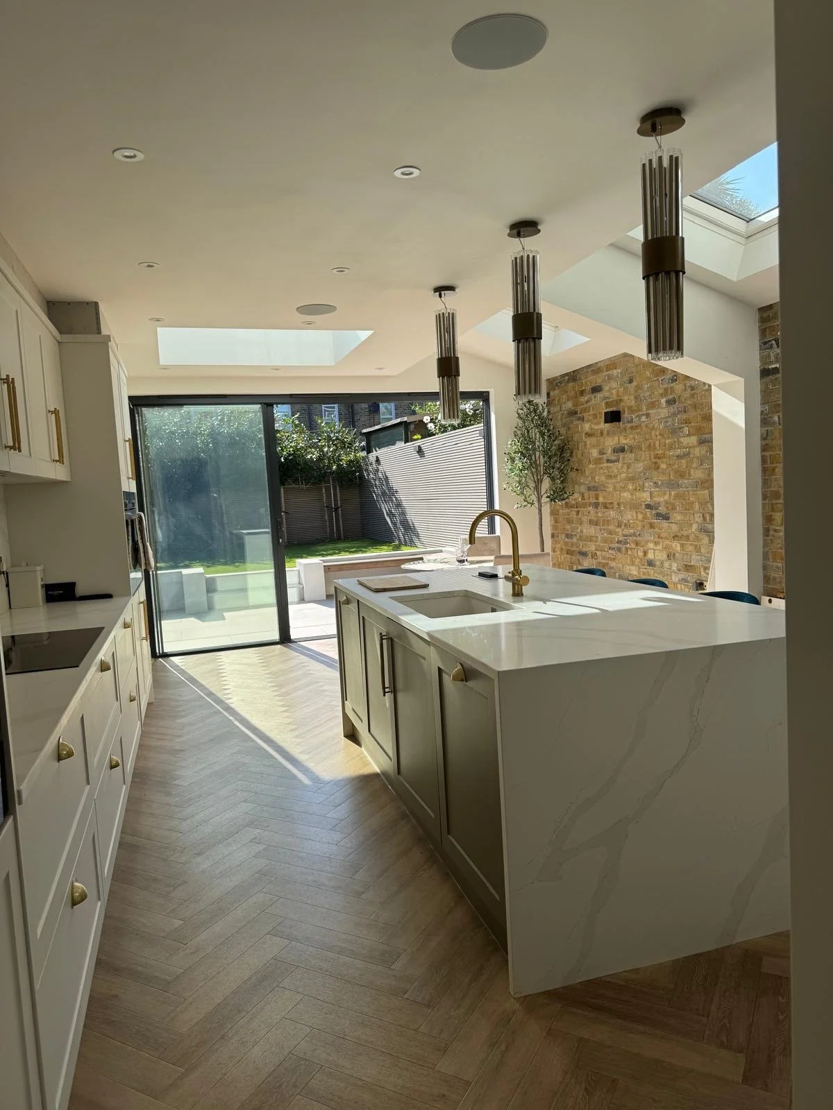 Modern kitchen with white cabinets, a large island with a marble countertop, brass fixtures, pendant lights, hardwood floors, and sliding glass doors leading to a backyard with lawn and fence.