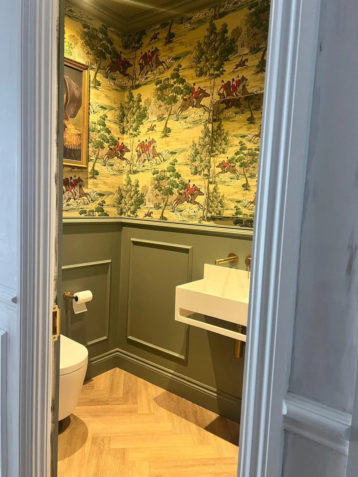 A small bathroom with green wainscoting, wallpaper with a scenic pattern of people riding horses, a white rectangular sink with brass fixtures, a white toilet, and wooden flooring.