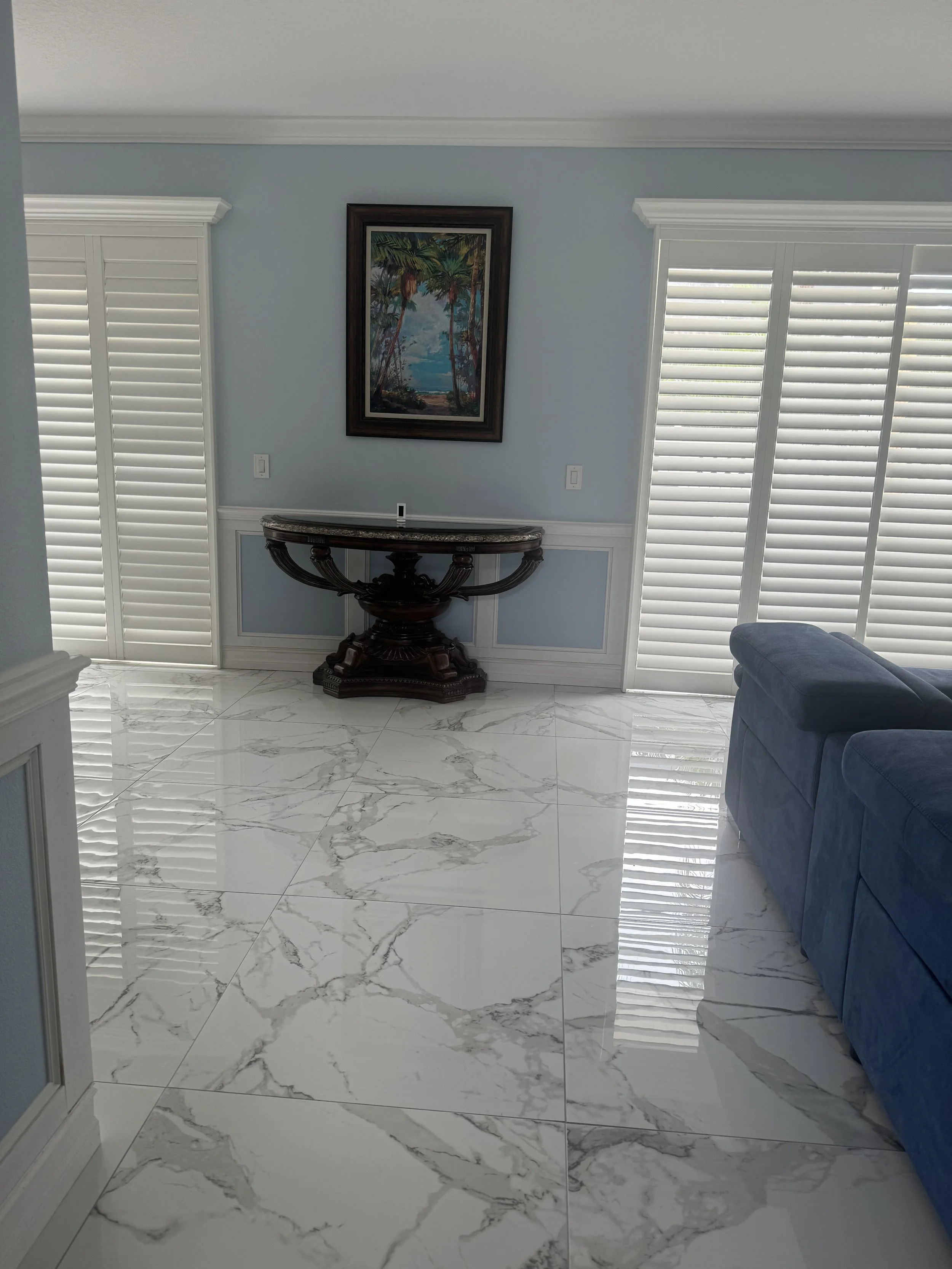 Living room with white marble tile flooring, a blue sofa, window shutters, a dark wood decorative table against the wall, a framed tropical landscape painting, and light blue walls.