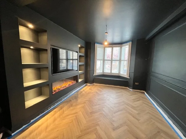 Empty living room with dark walls, built-in shelves, a wall-mounted TV, an electric fireplace, and a large window with blinds.