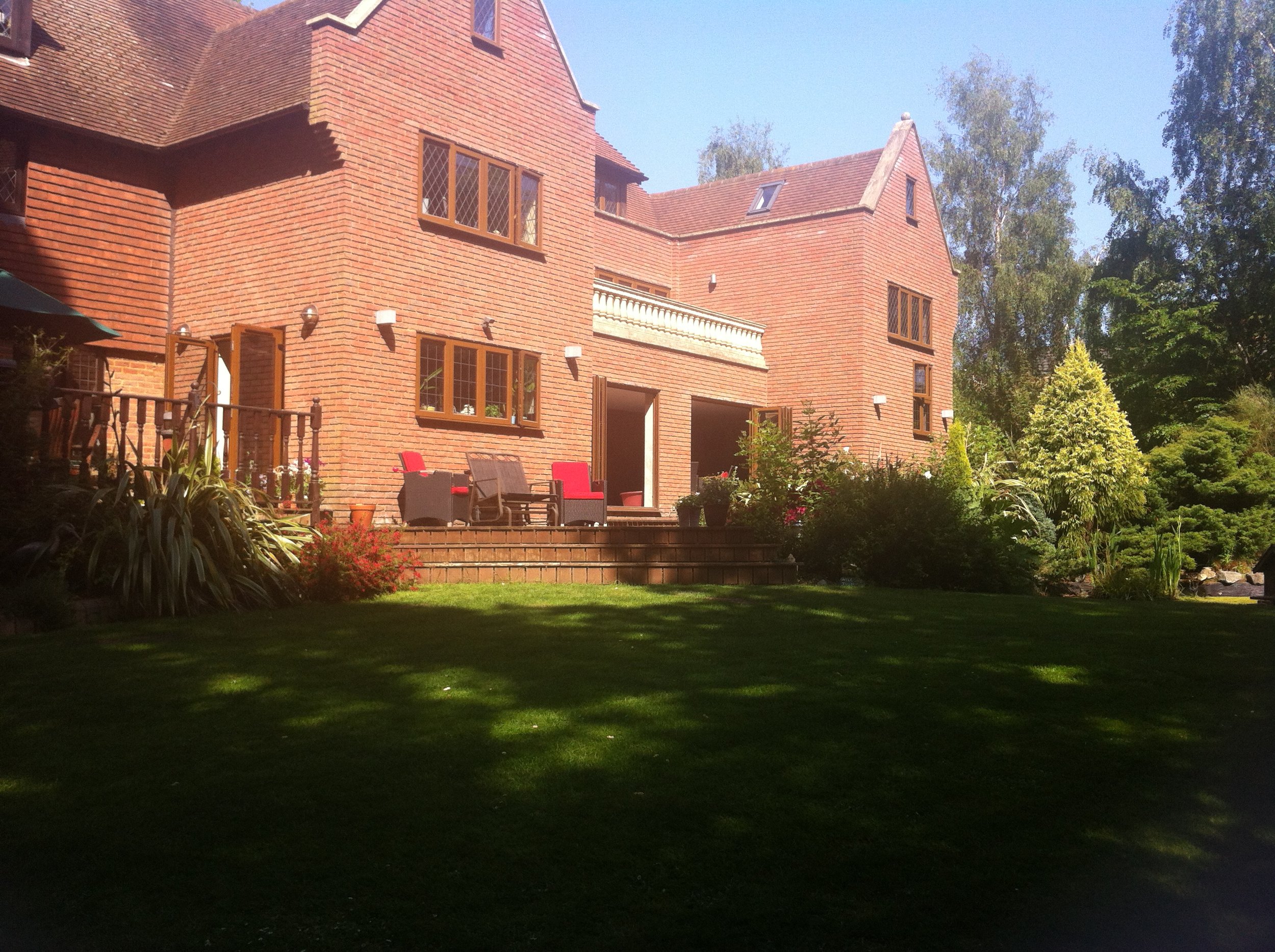 A large red brick house with a multi-level patio and outdoor furniture, surrounded by well-maintained garden and trees on a sunny day.