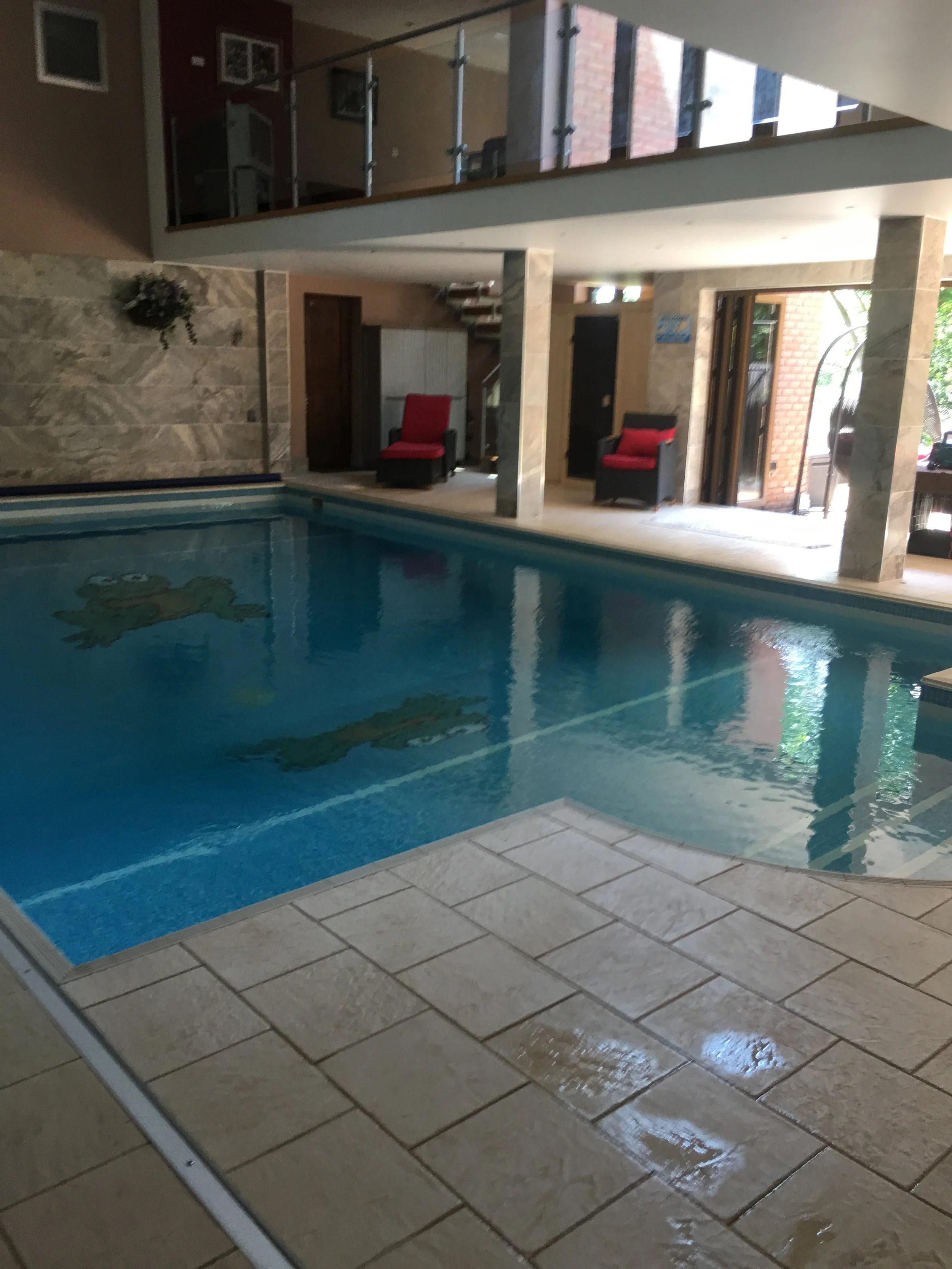 Indoor swimming pool with tiled floor and walls, pool with Elmo character design, and seating area with red cushioned chairs.
