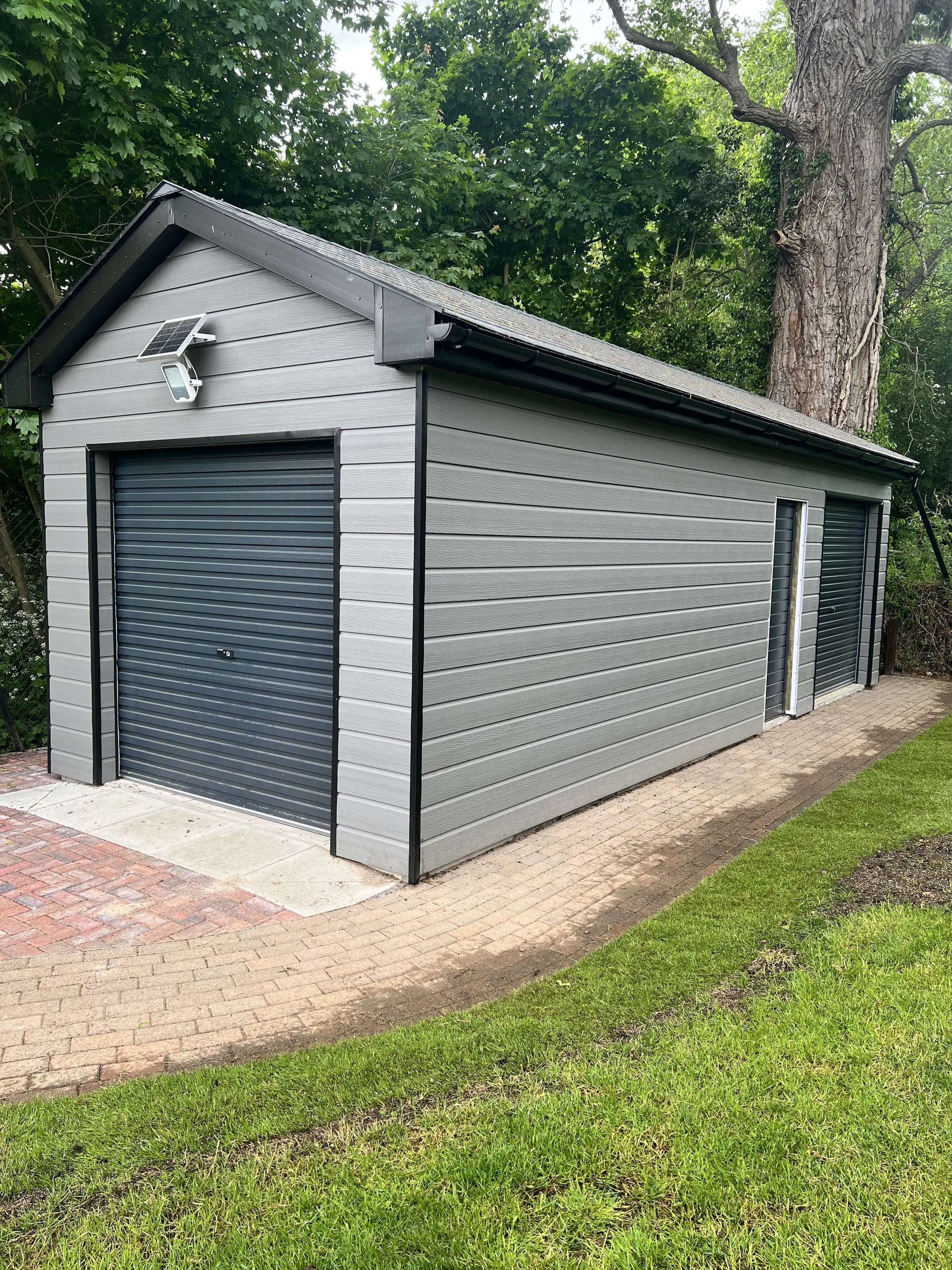 Gray metal shed with black rolling doors, situated on a brick walkway, with a grassy area and trees in the background, and a security light mounted above.