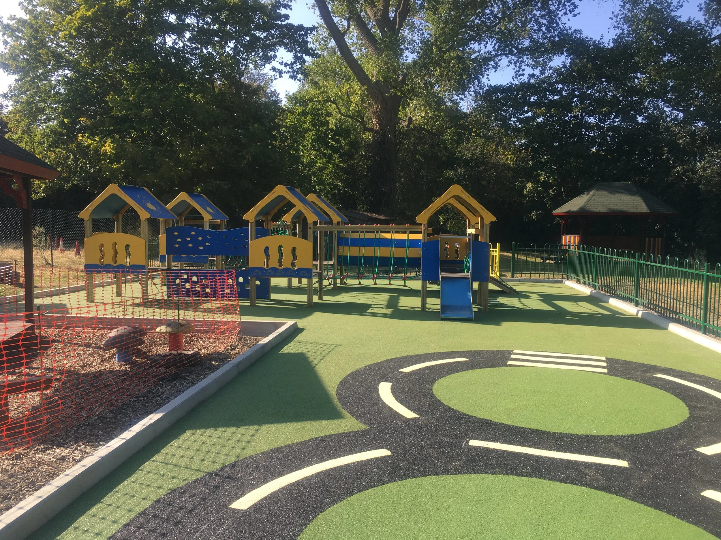 Colorful playground with a small blue slide, play structures with yellow roofs, a fenced area, and a painted course on the ground, surrounded by trees on a sunny day.