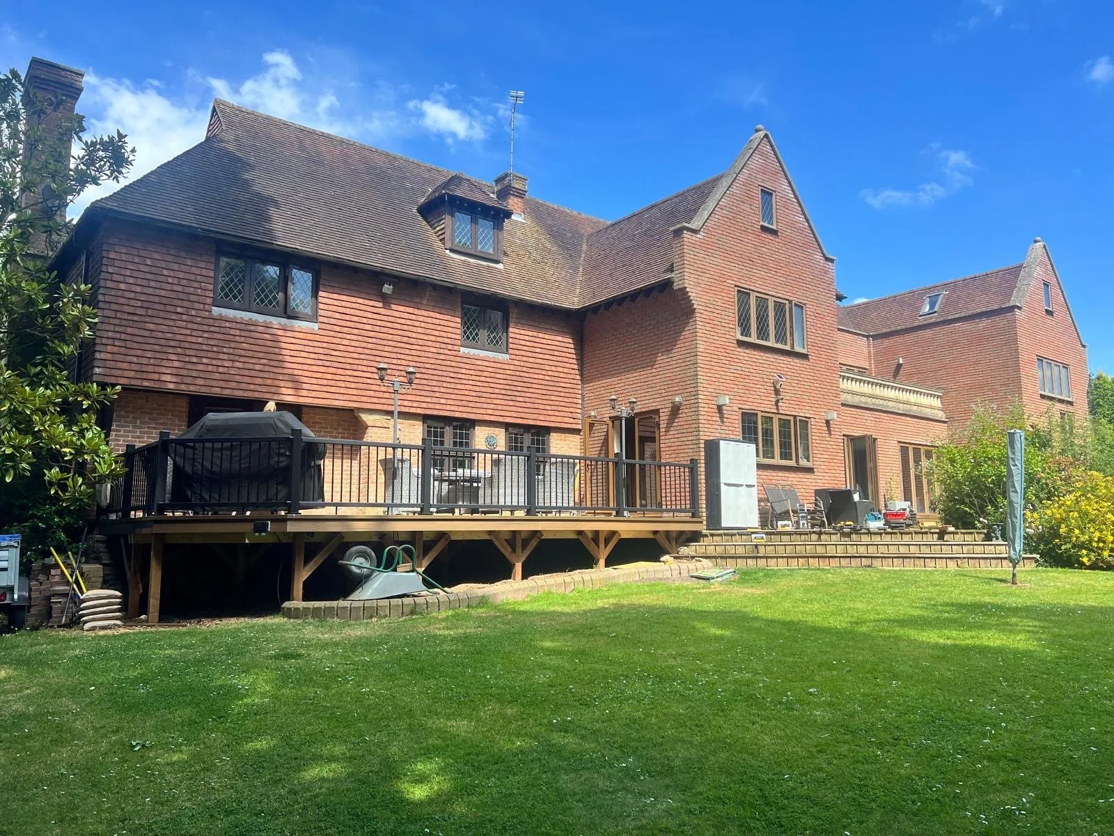 Large brick house with multiple gabled roofs and a wooden deck in the backyard, under blue sky with clouds.