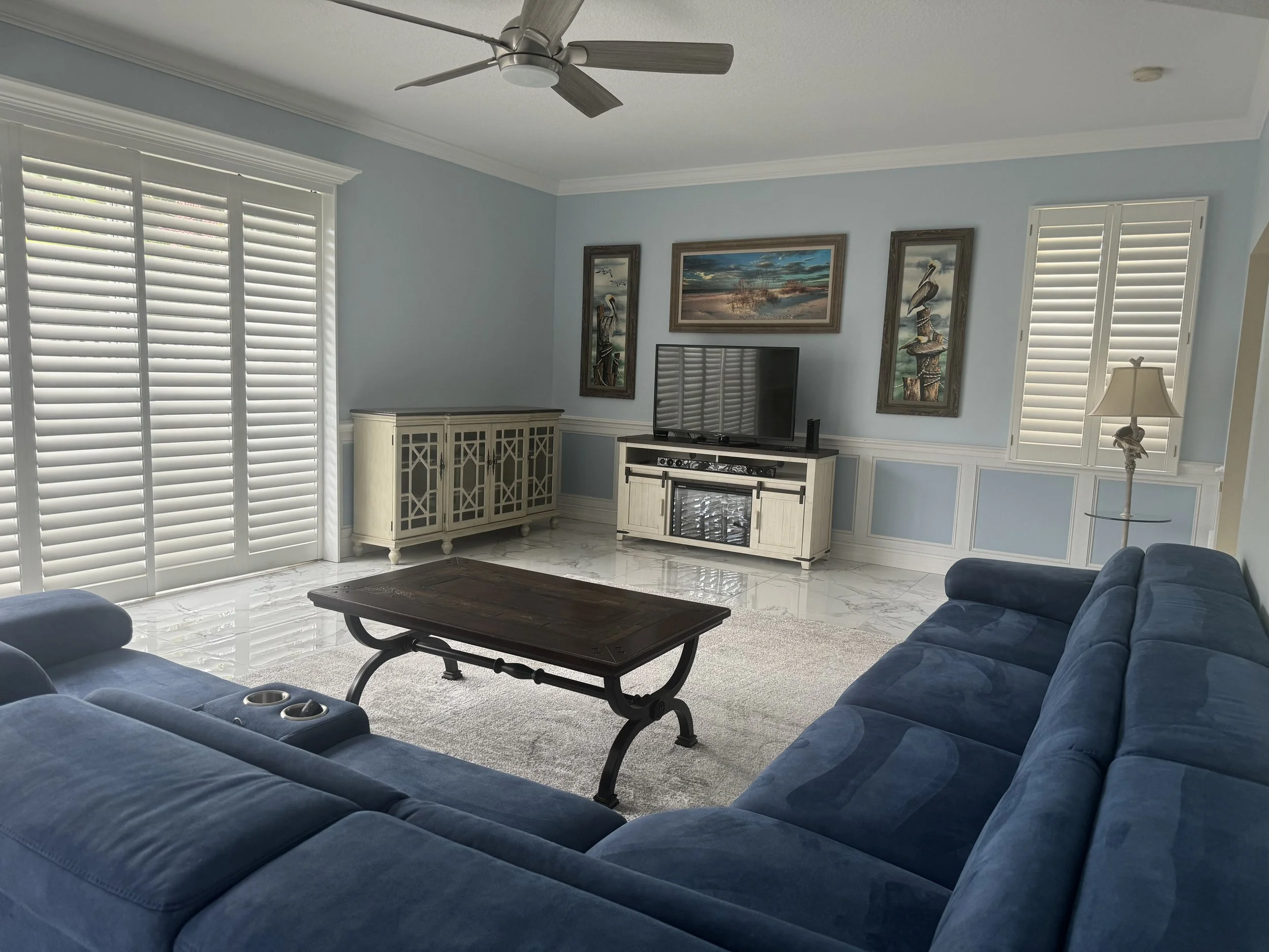 Living room with light blue walls, white window shutters, a large blue sectional sofa, a dark wooden coffee table, and a TV on a white stand. There's a ceiling fan and framed artwork on the wall.