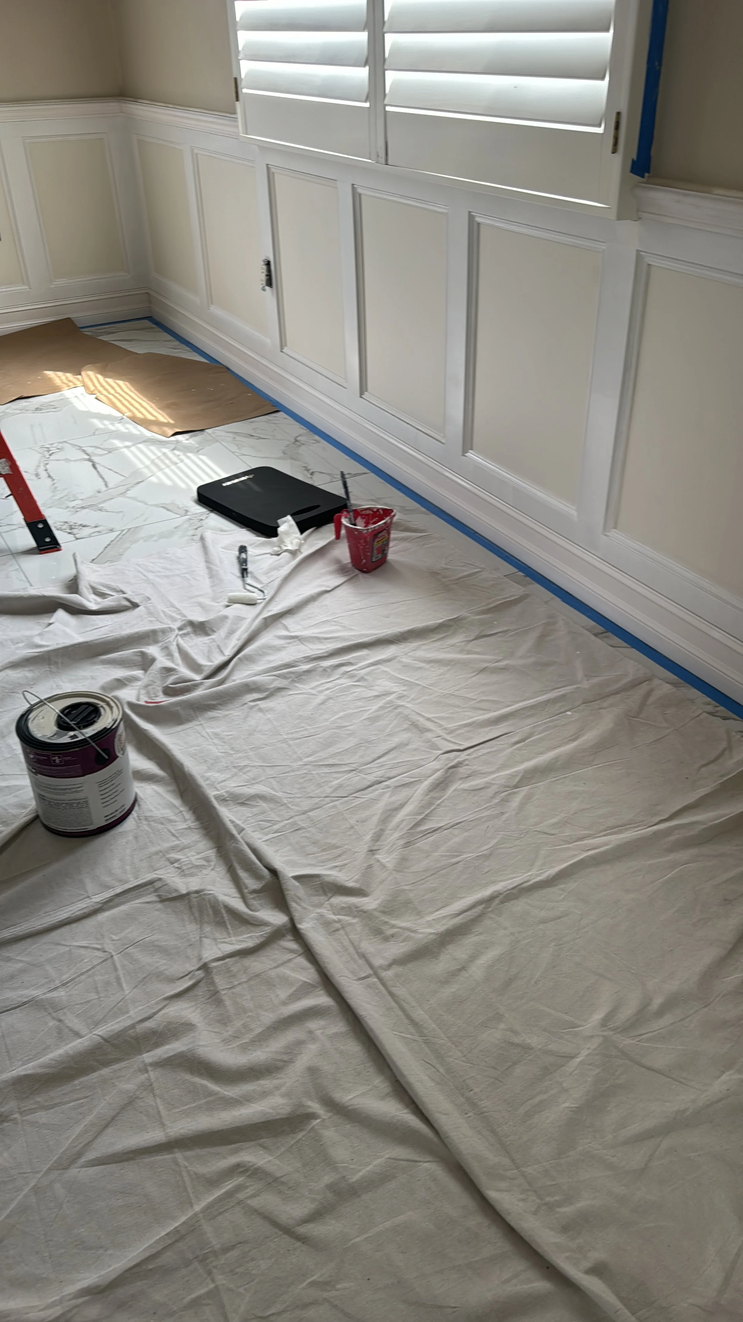 A room undergoing painting with protective plastic covering the floor, paint supplies, brushes, and buckets, and wainscoting on the walls.