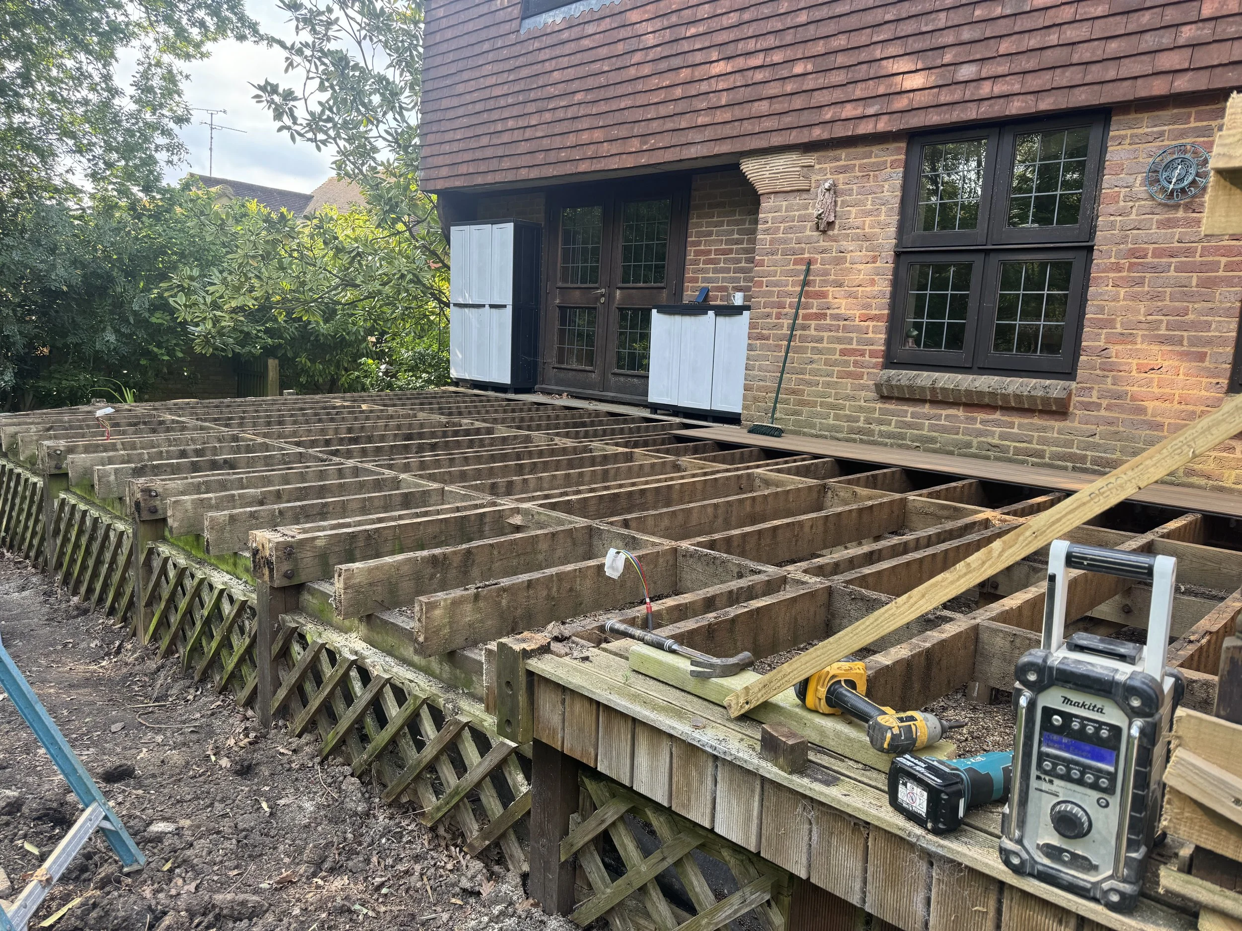 Wooden deck construction in progress outside a brick house with black-framed windows. Tools and materials like a drill, measuring tape, and lumber are on the deck. Some blue and white window shutters are mounted on the house.