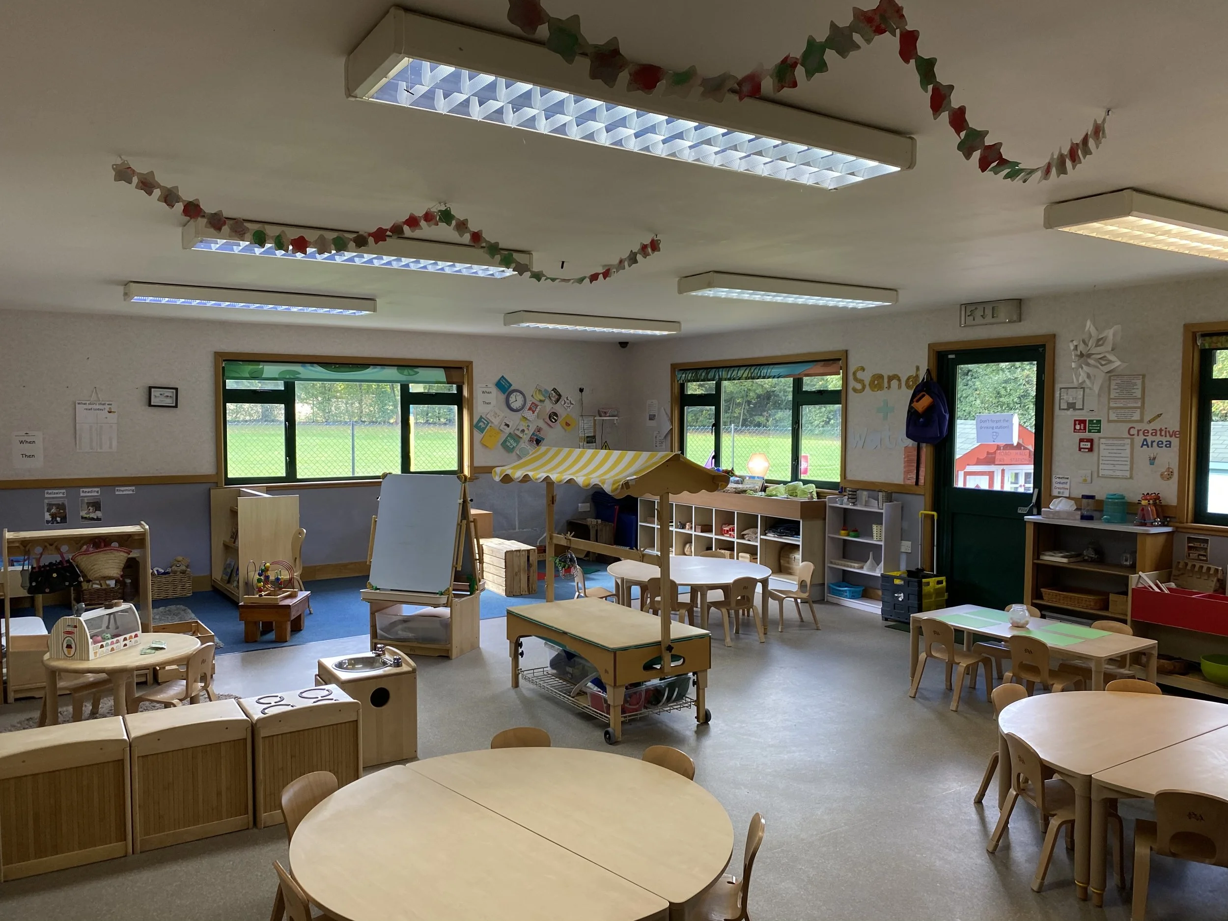 Empty preschool classroom with wooden tables and chairs, educational toys, storage shelves, decorated walls, and large windows showing greenery outside.