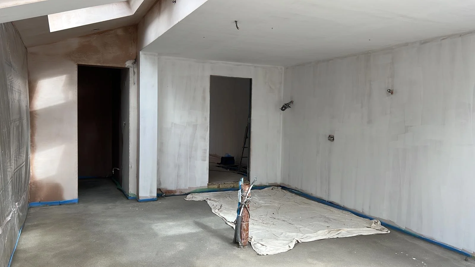 Interior of a room under construction with unfinished white walls, a doorway, exposed wiring, and a blue protective covering along the floor edges.