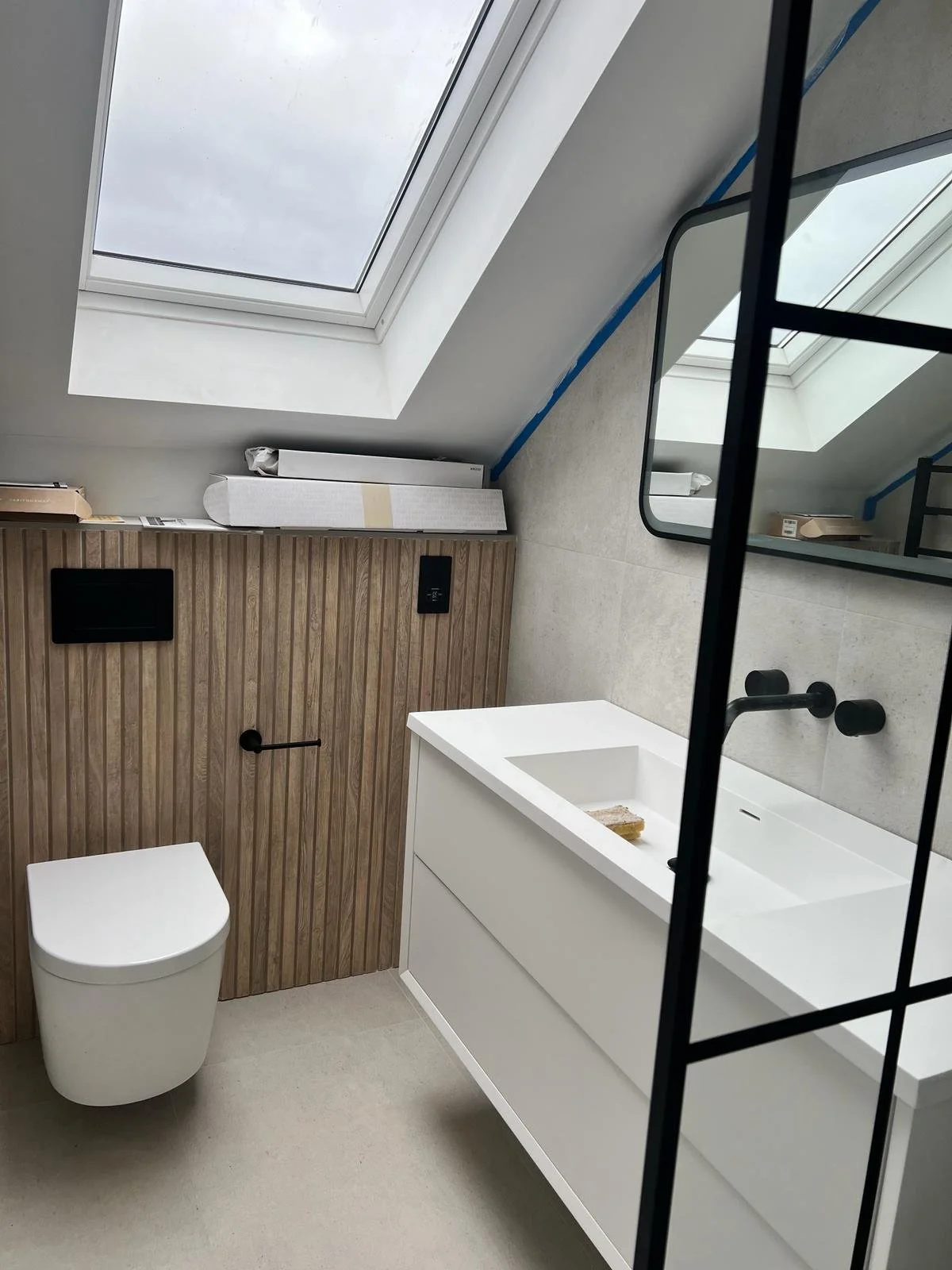 Modern bathroom with skylight, white vanity with integrated sink, black fixtures, wooden panel wall, mirror, and toilet, under a sloped ceiling.
