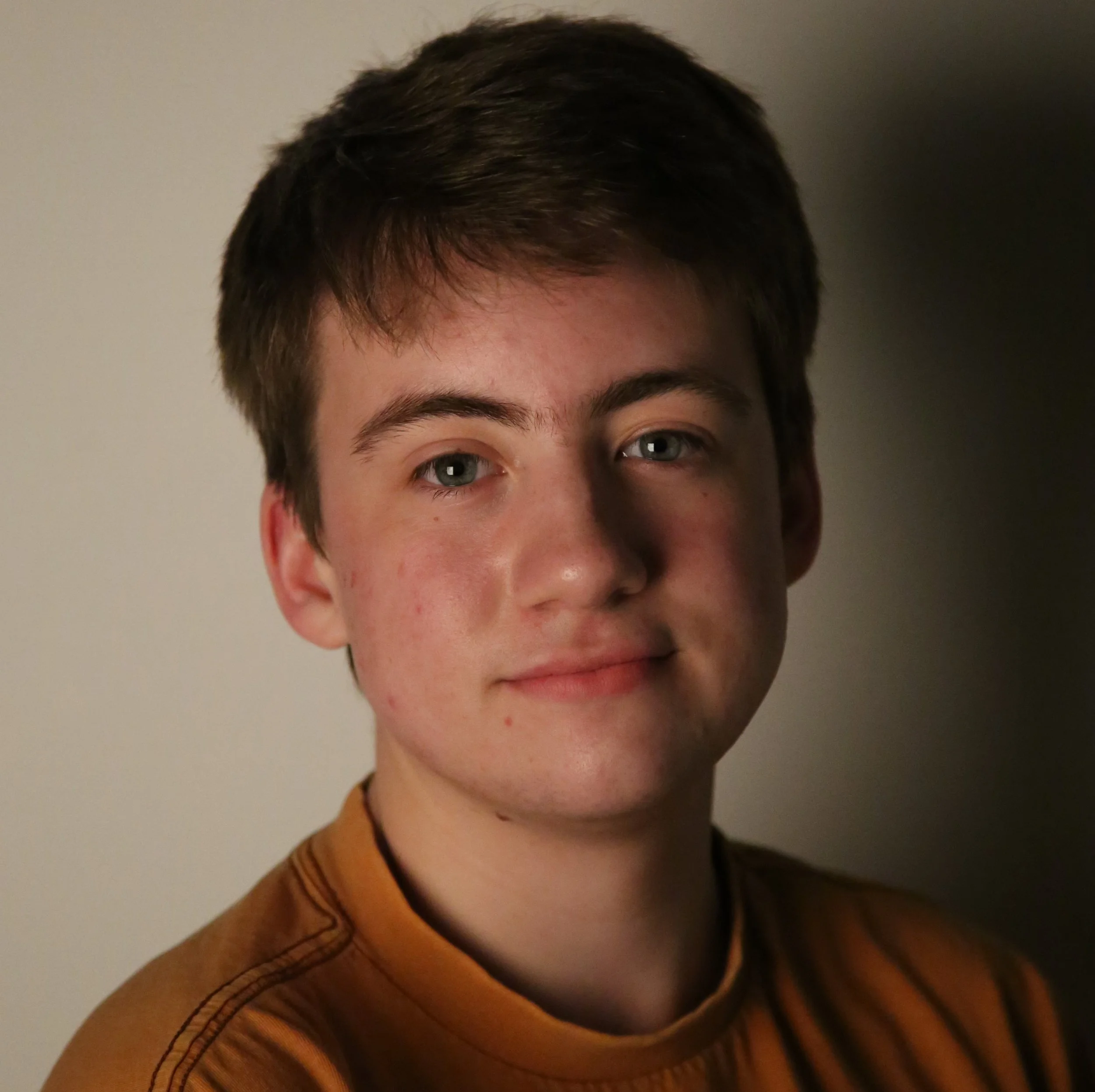 Headshot of a young man in his early 20s. He has short brown hair, blue eyes, and fair skin. He is wearing a neutral orange T-shirt.