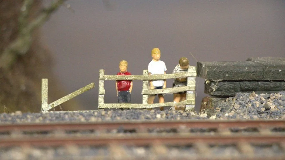 Britain's Biggest Model Railway BBMR , Heaton Lodge Junction, showing Simon George and two friends as three miniature figurines on a model train platform with train tracks in the foreground.
