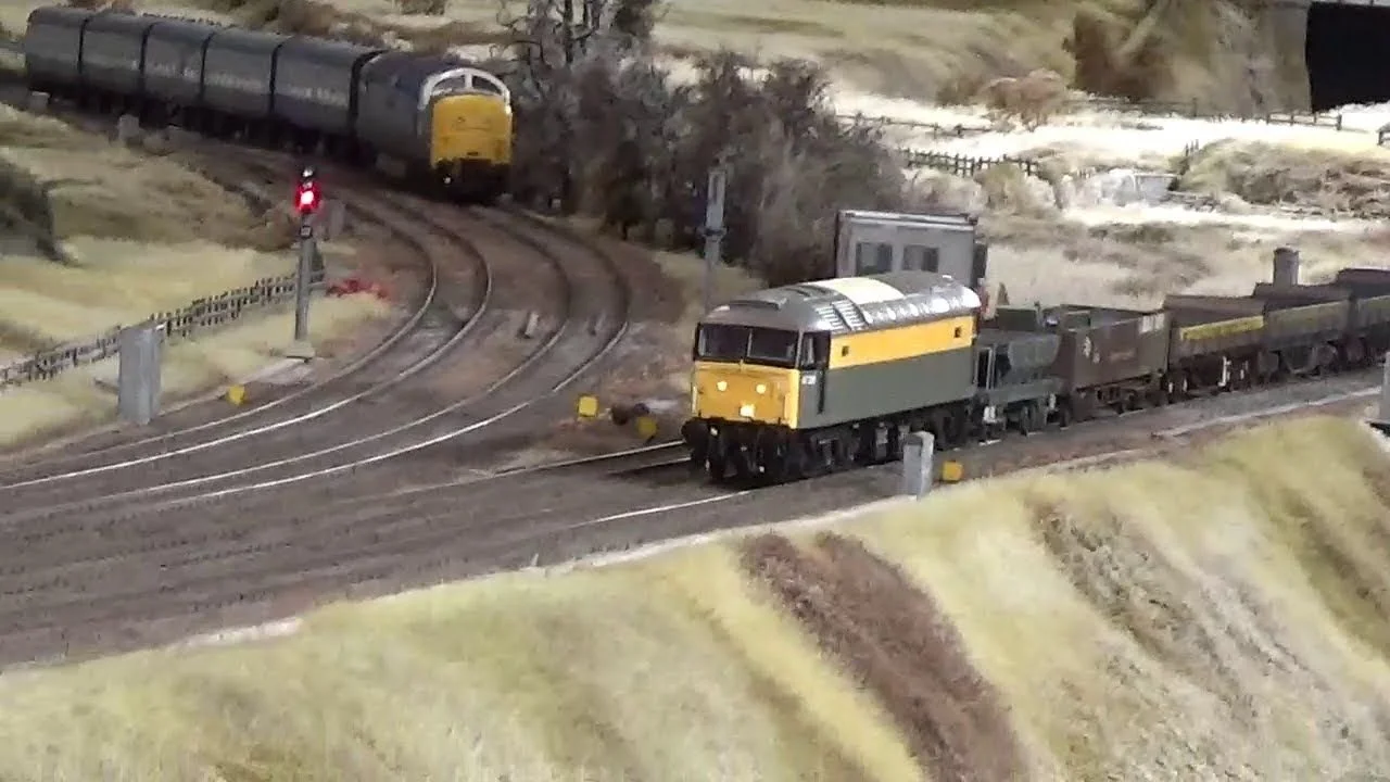 Model train scene with a locomotive and freight cars on track, with a signal showing red light, and a landscape with trees and hills in the background.