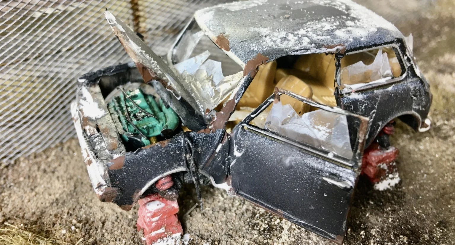 Simon George can be seen here modelling in action, on his Heaton Lodge Junction, known as Britain's Biggest Model Railway, we see a black car heavily damaged, with exposed interior, red brick-like supports, and debris around it on dirt ground.