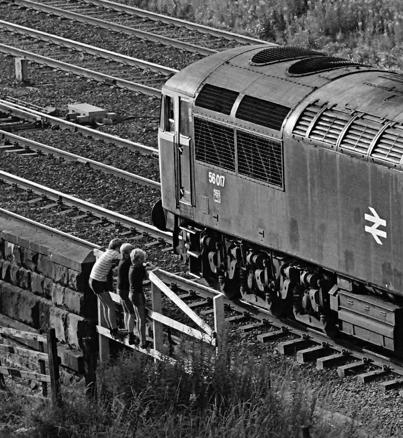 Britain's Biggest Model Railway BBMR , Heaton Lodge Junction, showing Simon George and a friend observing the trains on a small wooden bridge over train tracks, looking at an approaching locomotive train in black and white.