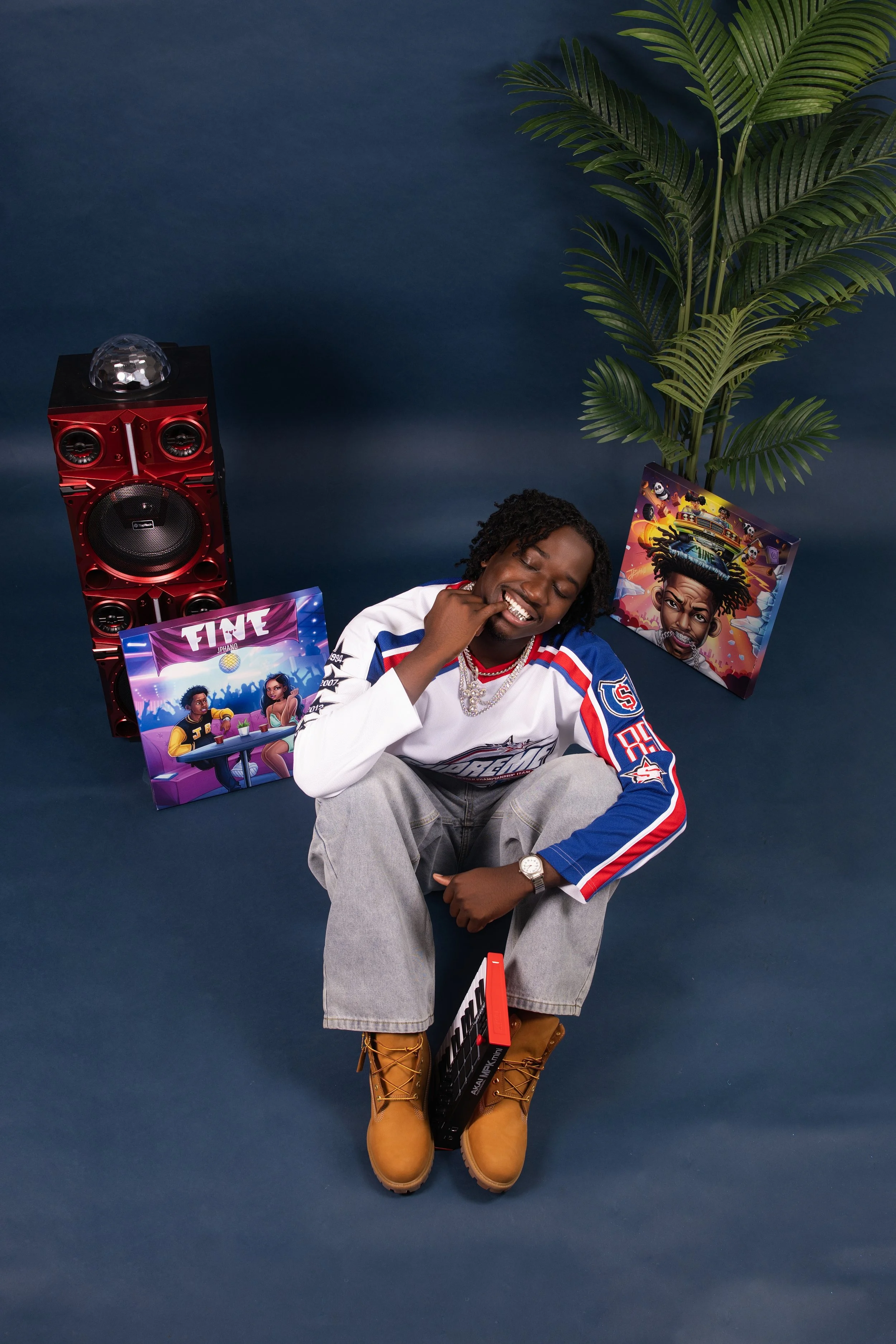 A young man sitting on the floor, smiling with his eyes closed, wearing a white, blue, and red sports jersey, gray pants, and tan boots. Surrounding him are colorful posters, a large red speaker, and a potted plant against a dark blue background.
