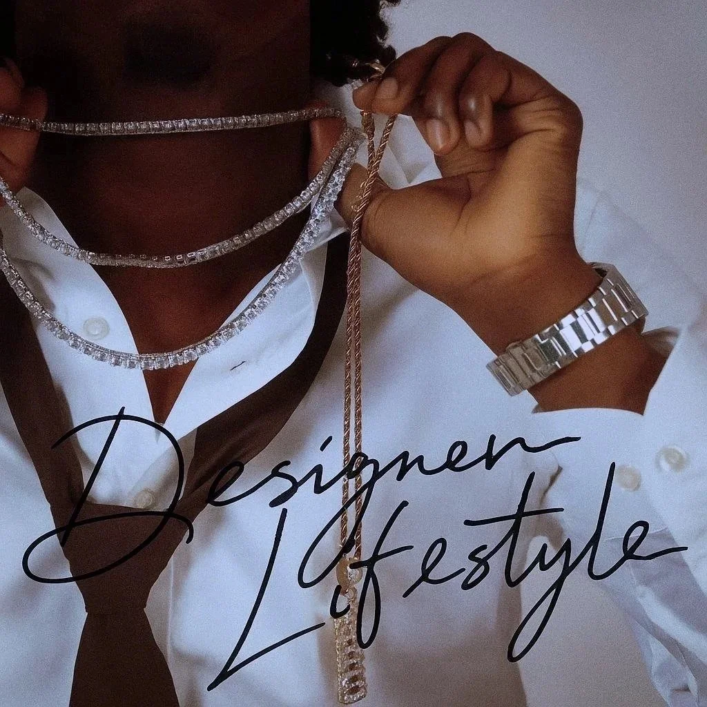 A person holding a gold chain necklace near their neck, wearing a white shirt with a dark tie, a silver watch, and multiple diamond necklaces. The image has the text "Designer Lifestyle" written in cursive.