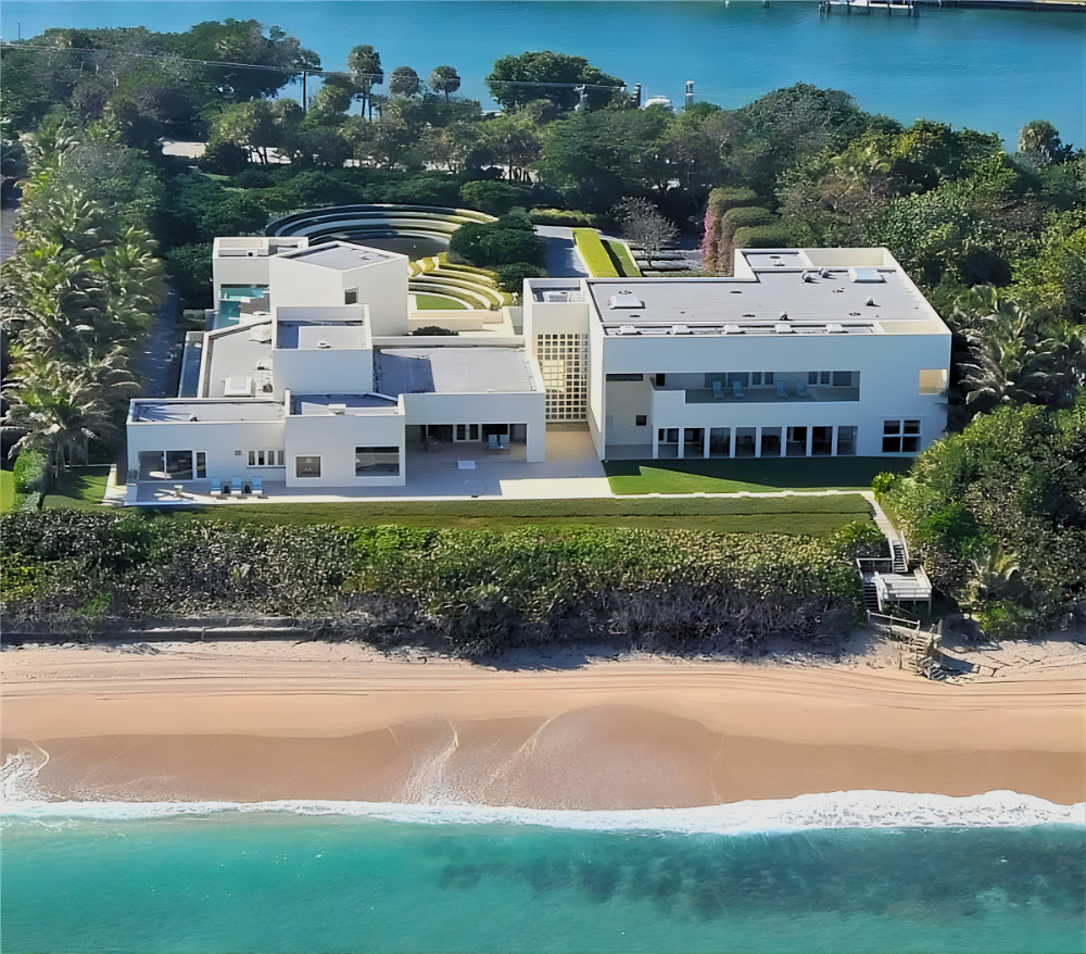 Aerial view of a modern white mansion on a beach, with a lush green yard, trees, and a body of water in the background.