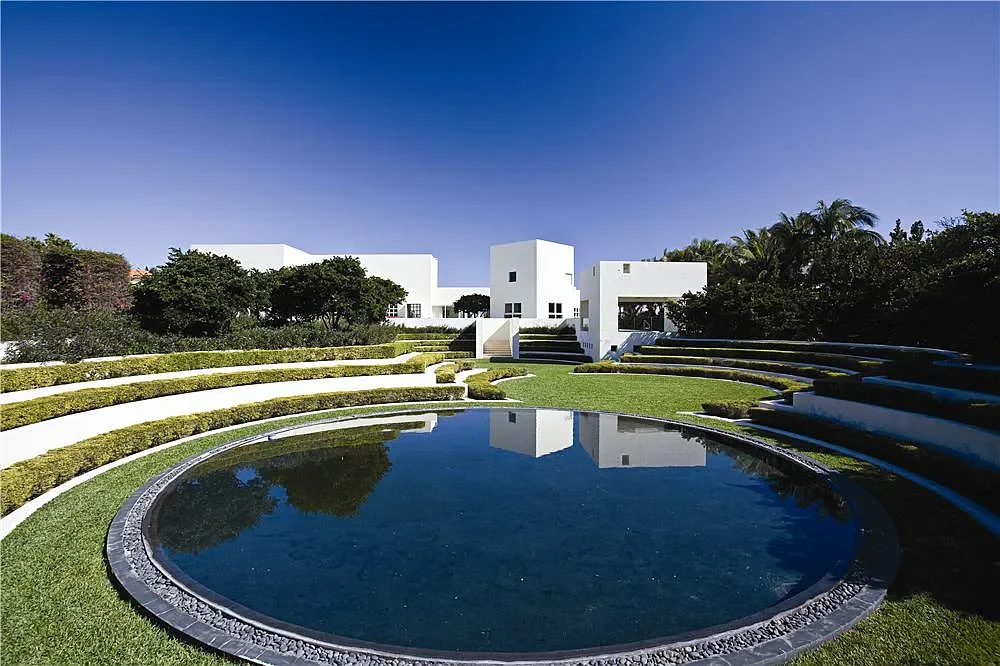 Modern white buildings with tiered gardens around a circular reflective water feature on a sunny day.