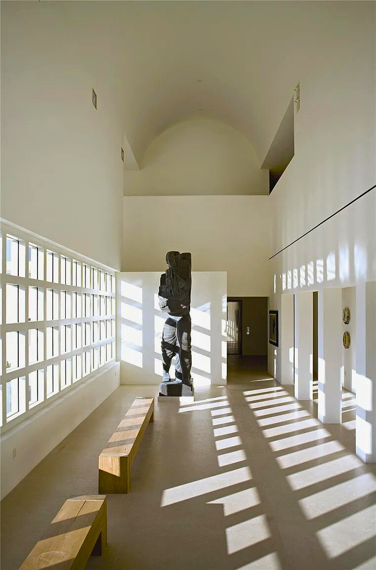 Interior of a modern art gallery with a wooden bench, a sculpture, and natural light casting shadows through windows.