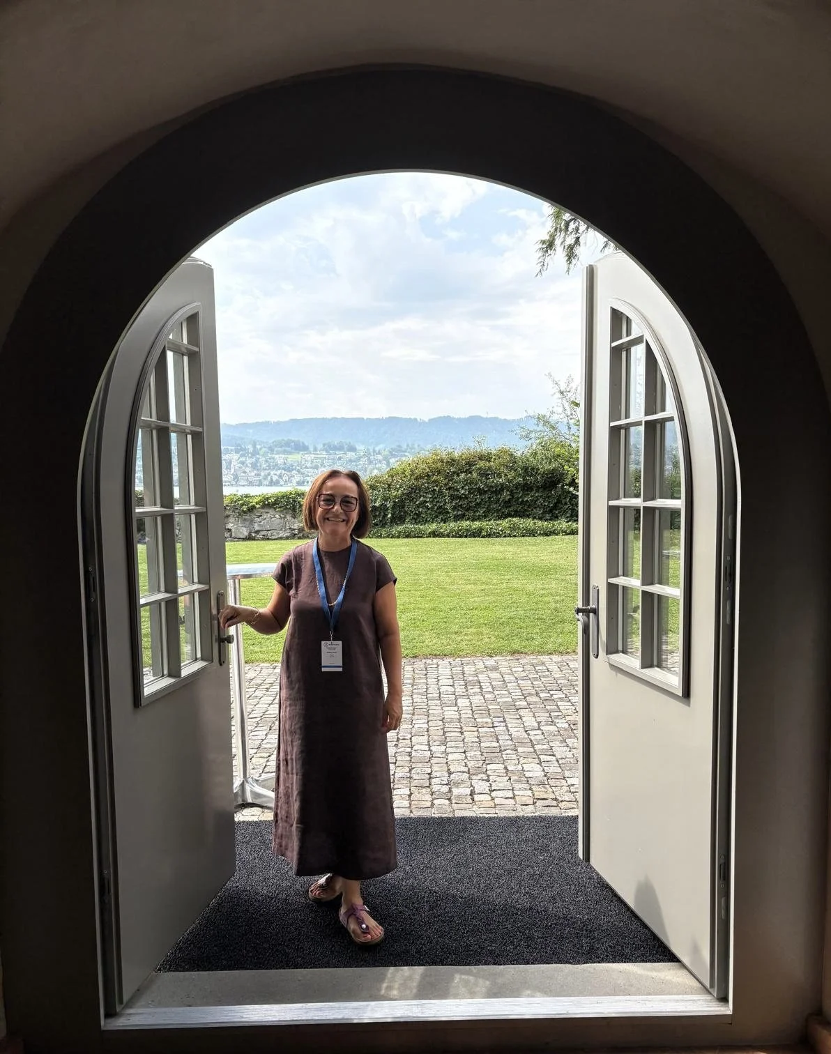 Woman standing in an open doorway outdoors with a city, hills, and cloudy sky in the background.