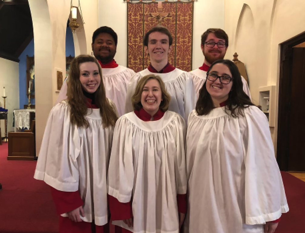 Group of seven choir members dressed in white robes with red accents, standing inside a church with wooden furnishings and religious decorations.