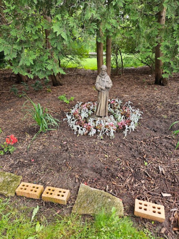 A garden scene featuring a religious statue of a saint surrounded by pink and white flowers, with trees in the background and some decorative bricks and stones in the foreground.