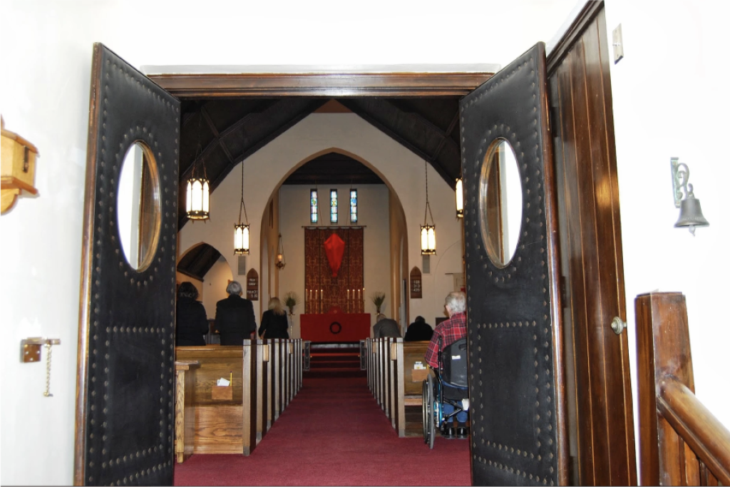View inside a church through open double doors showing pews with people, an altar, and religious decorations.