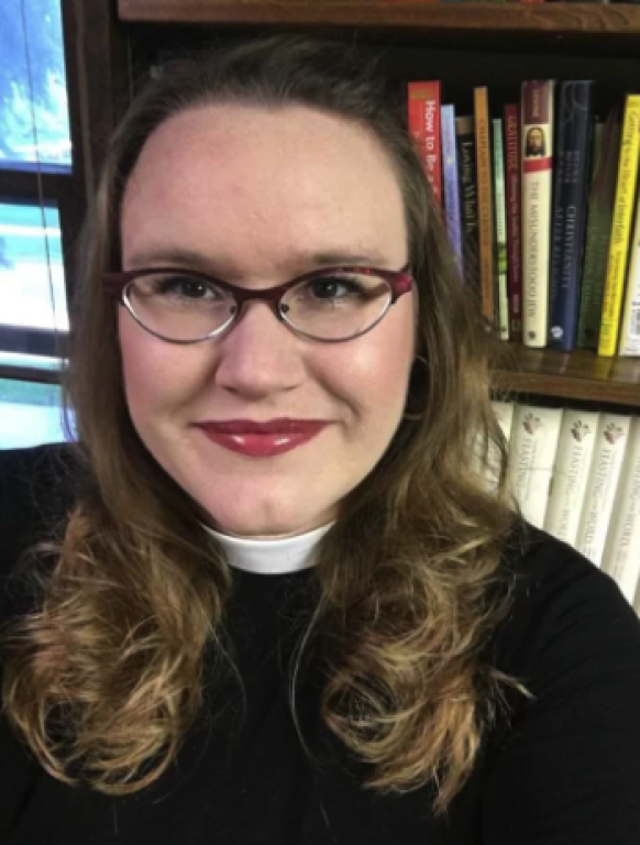 A woman with long, wavy hair wearing glasses and a black top with a white collar, smiling in front of a bookshelf filled with books.