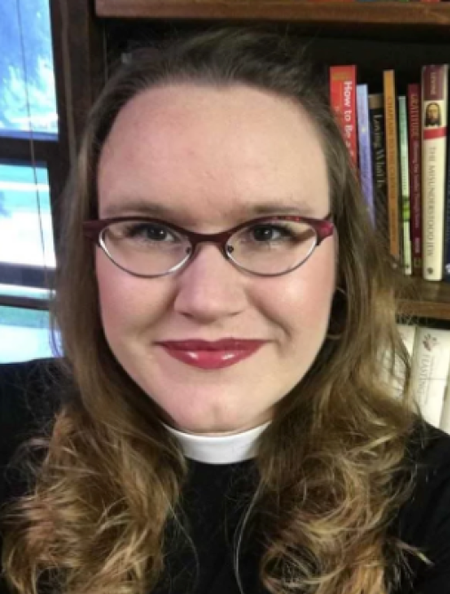 Close-up of a woman with long wavy hair, wearing glasses, smiling, in front of a bookshelf filled with books.